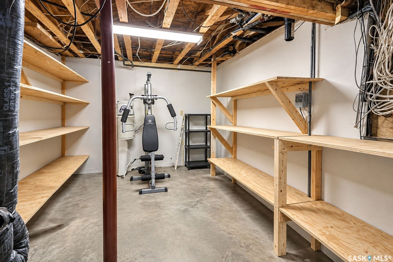 Basement area with concrete flooring, exposed ceiling joists, built-in wooden shelving, and a fluorescent light fixture