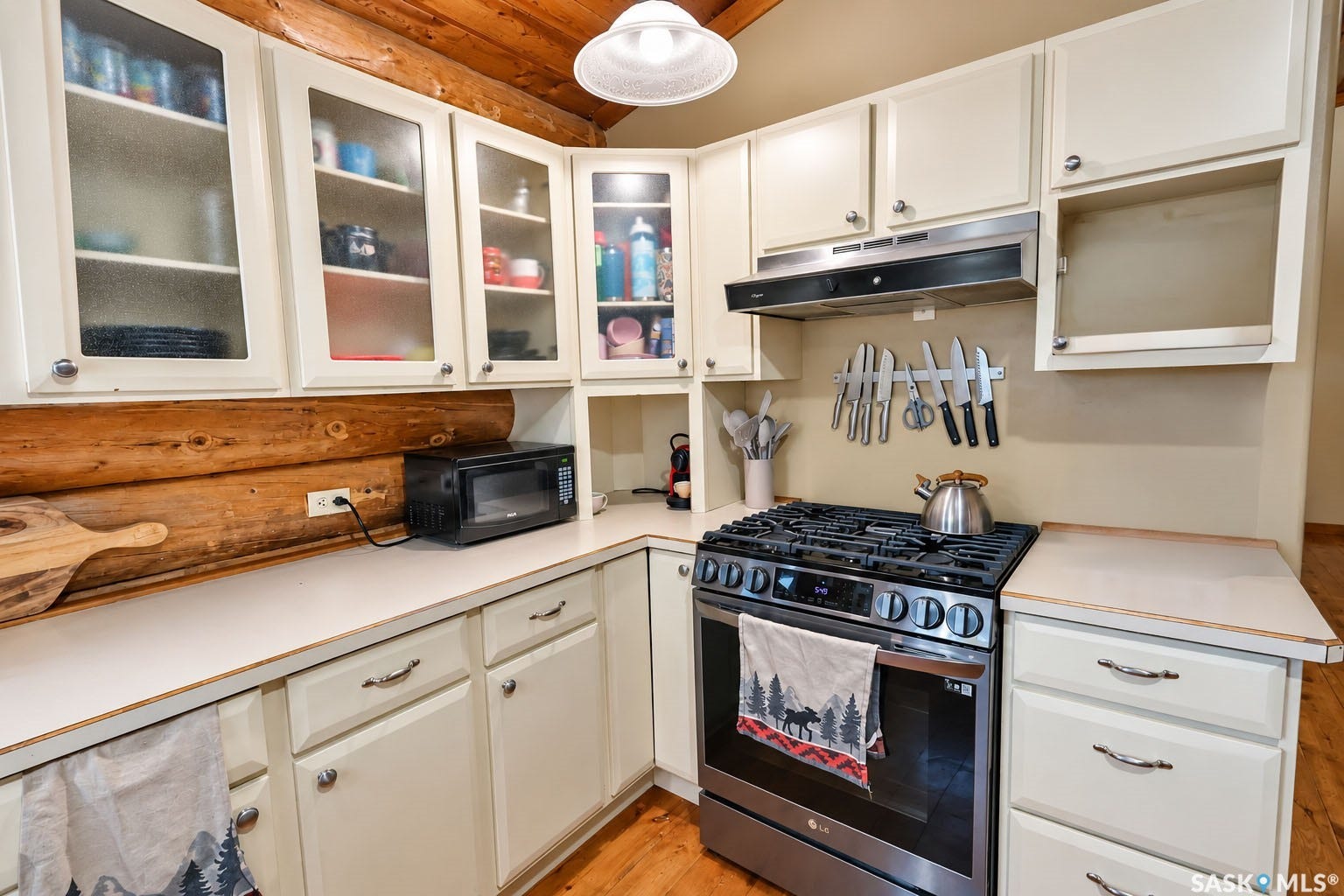 White cabinetry with glass-front upper doors, log wall detailing, gas range with stainless steel hood, and light-toned countertops