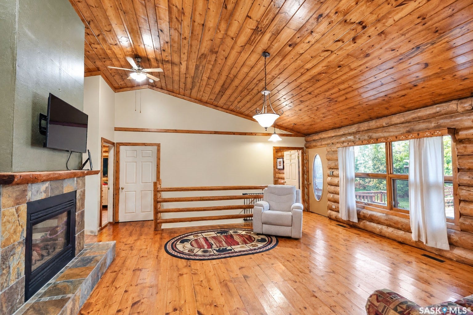 Tongue and groove wood-finish ceiling, hardwood flooring, stone-clad fireplace, log-style wall construction, and large window with exterior scenery
