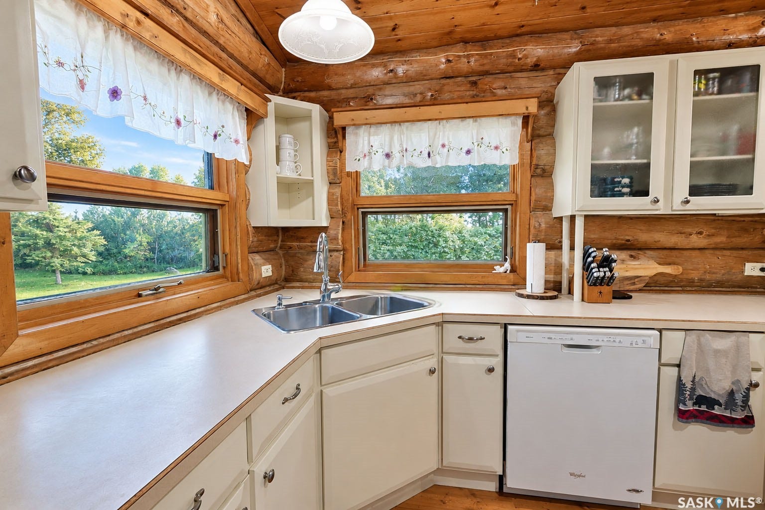 Wood-paneled interior walls, white cabinetry, corner double basin stainless steel sink, white countertops, and wood-frame windows
