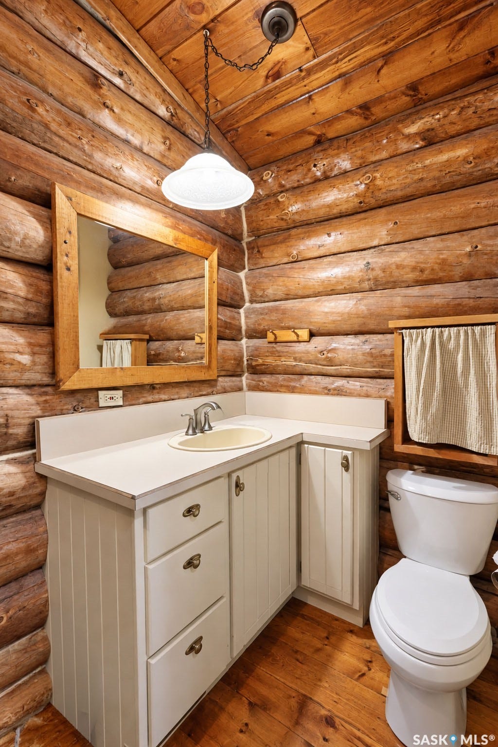 Log wall paneling, wood-finish flooring, white vanity with integrated sink, chrome-finish faucet, and a wall-mounted mirror