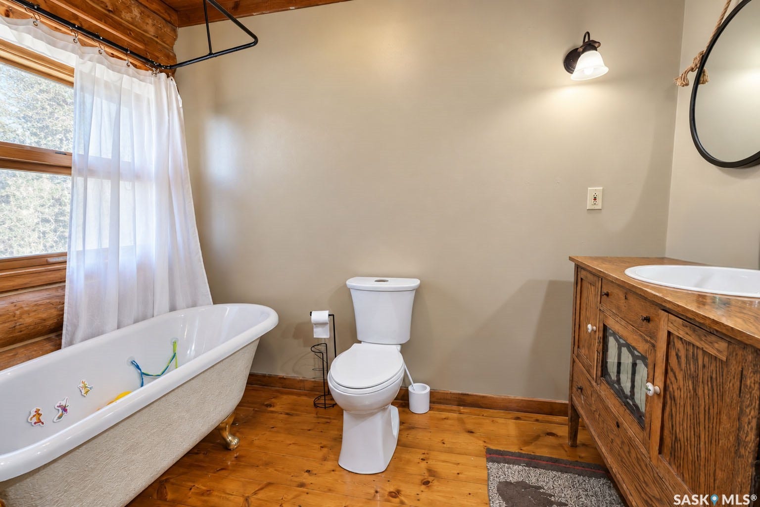 Freestanding clawfoot tub, wood-finish flooring, wood vanity with integrated sink, wood-framed window, and log wall accent