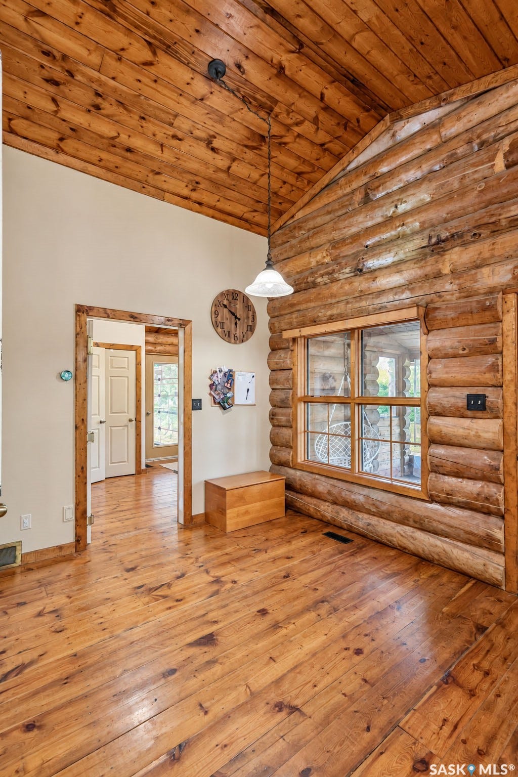 Vaulted wood-paneled ceiling with a hanging fixture, wood-plank flooring, and log wall construction with a window