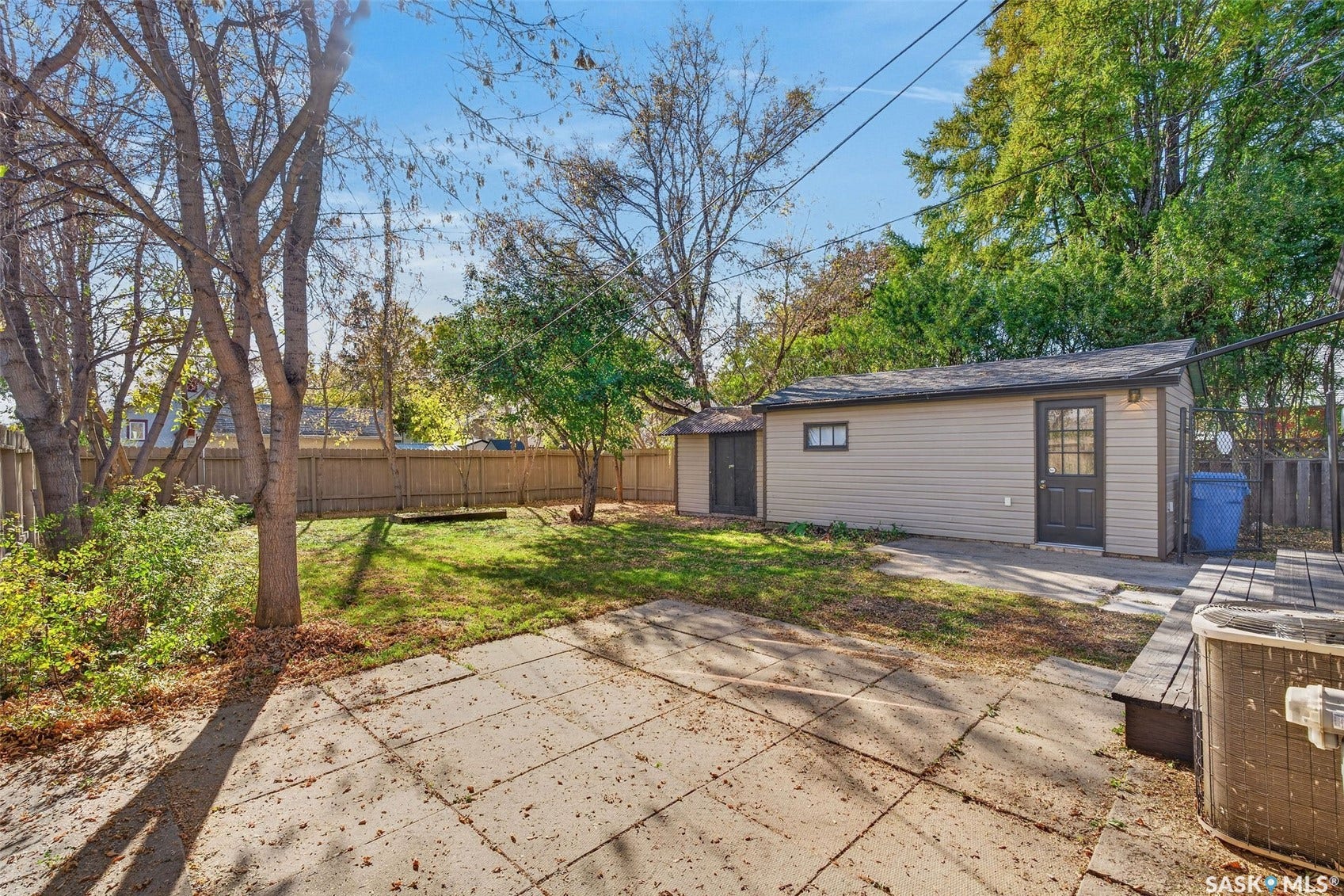 Expansive backyard featuring a concrete patio, grassy area, mature trees, and a detached structure with siding and a shingled roof