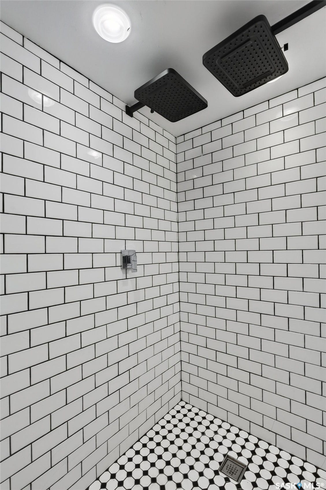 White subway tile shower walls, dual matte black rain shower heads, black and white hexagon mosaic tile flooring, and a square shower drain