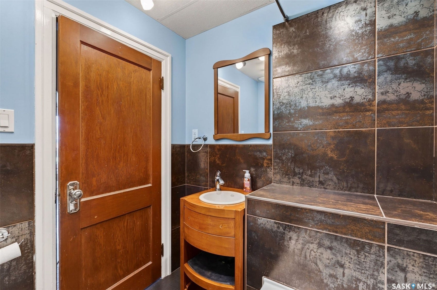 Wood door with panel detailing, wood vanity with integrated sink, wood-framed mirror, and tiled shower with built-in bench