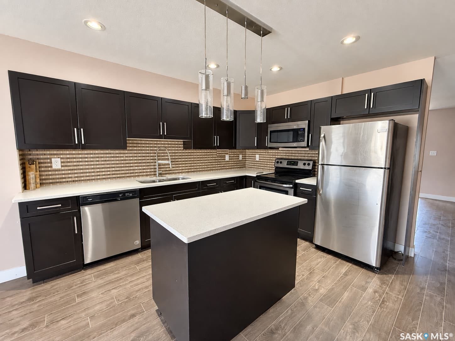 Kitchen featuring dark wood-finish cabinetry, stainless steel appliances, a center island with a light-toned countertop, a linear pendant light fixture, and wood-look tile flooring