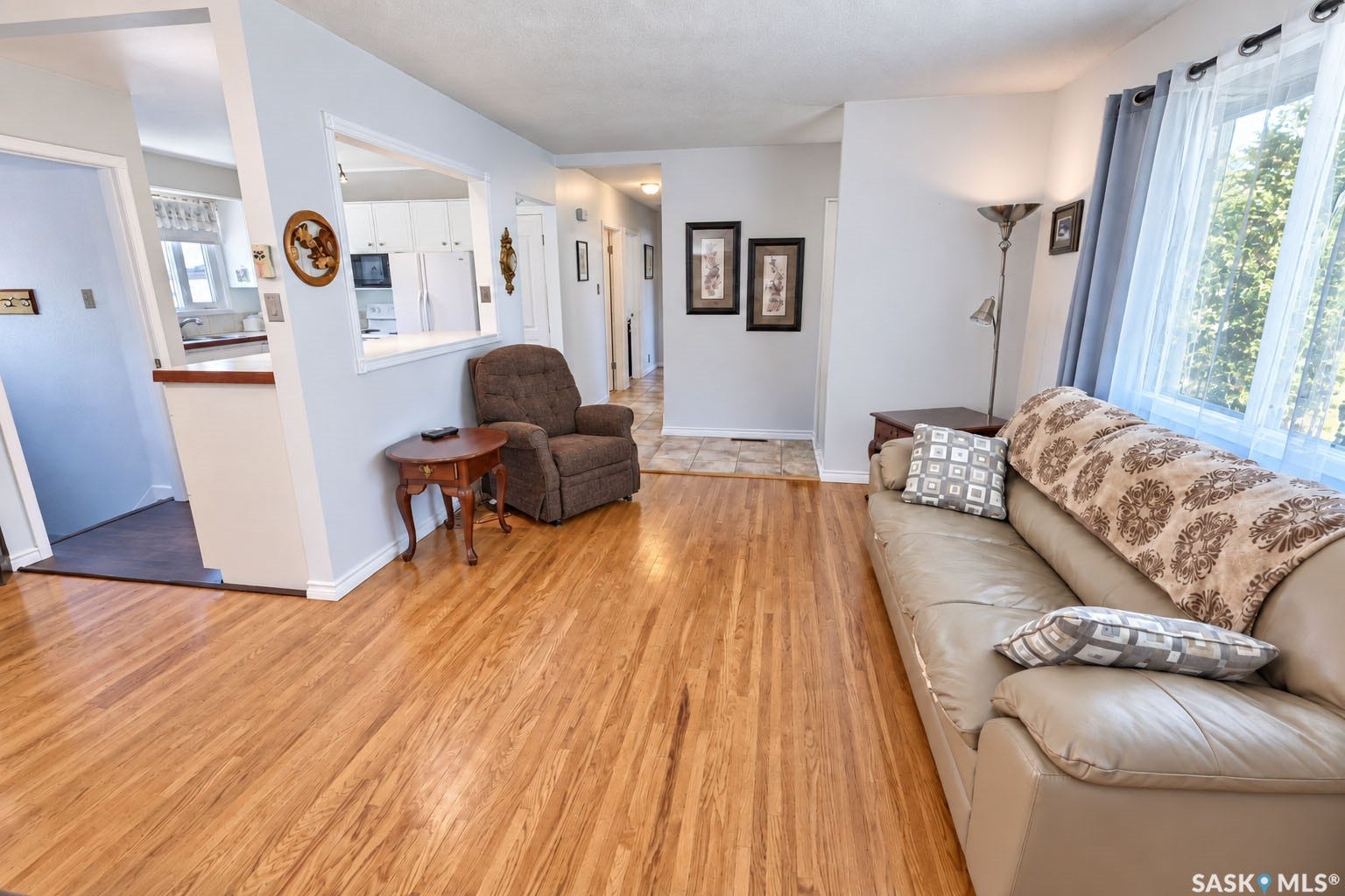 Hardwood flooring extending throughout, window with white sheers and blue curtains, white baseboards, and a pass-through opening to the kitchen
