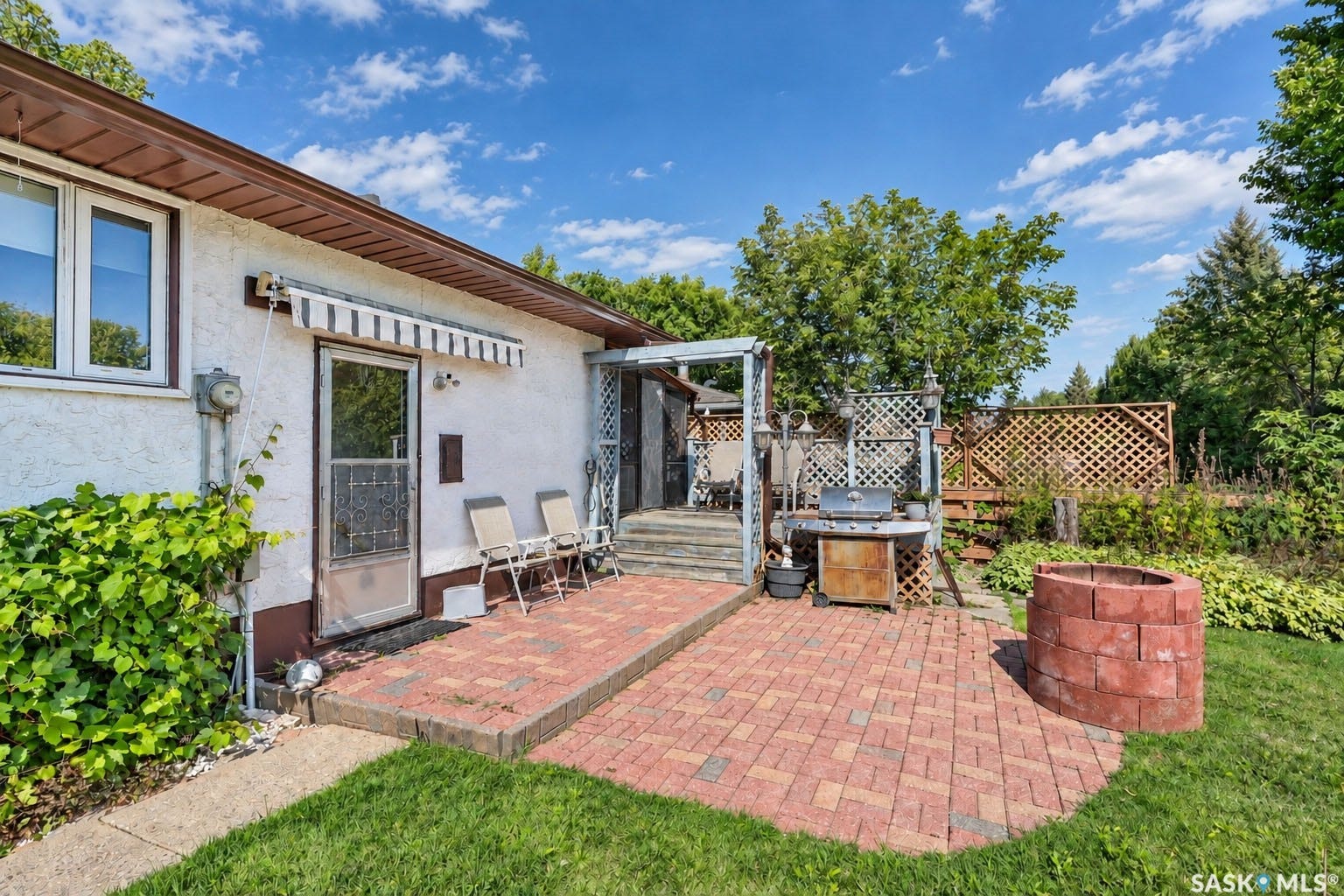 Brick paver patio with a built-in fire pit, leading to a raised wooden deck with lattice fencing