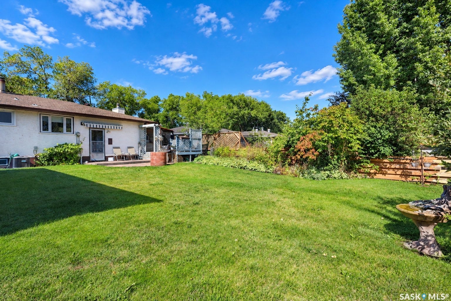 Expansive green lawn with mature trees, a ground-level patio, and a raised wooden deck
