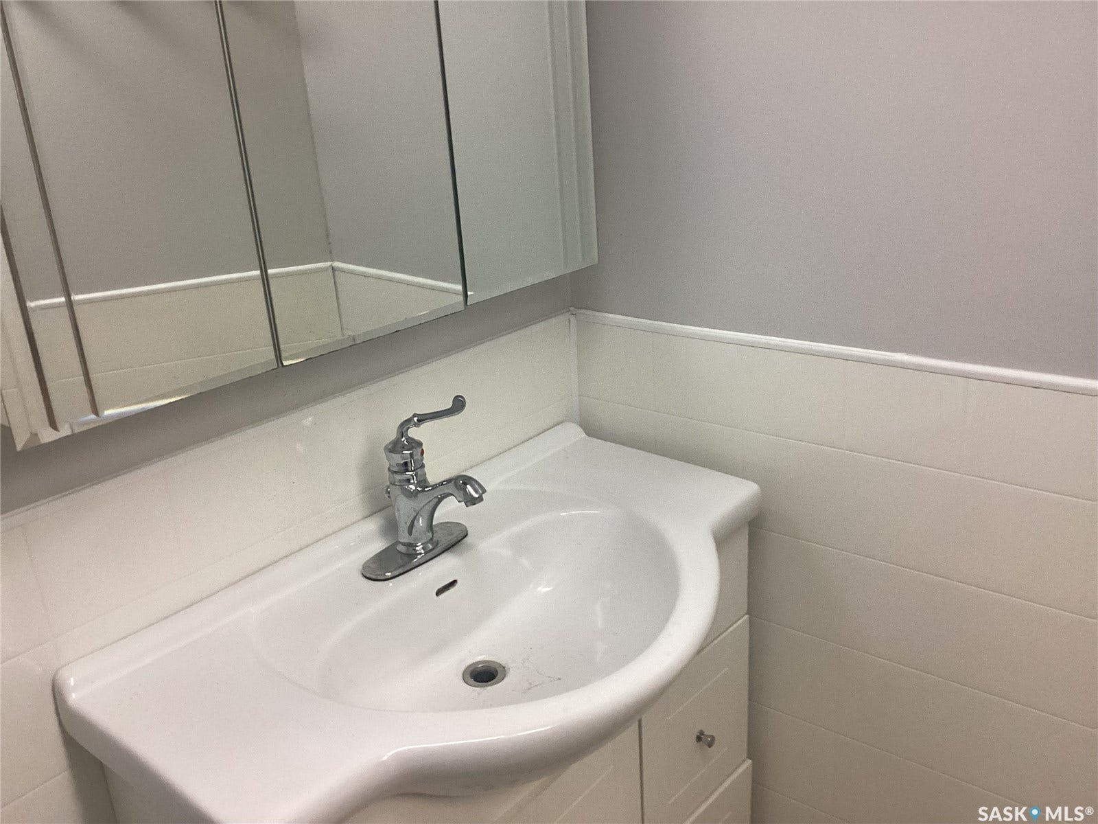 White vanity with a ceramic basin, chrome faucet, white paneled lower wall, and a mirrored medicine cabinet