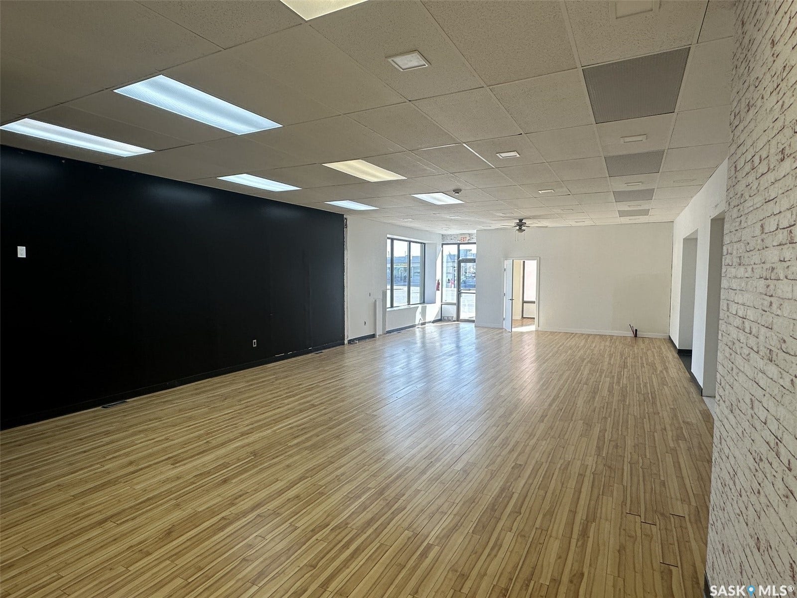 Expansive commercial space featuring wood-finish flooring, drop ceiling with integrated lighting, a black accent wall, and a textured brick-finish wall