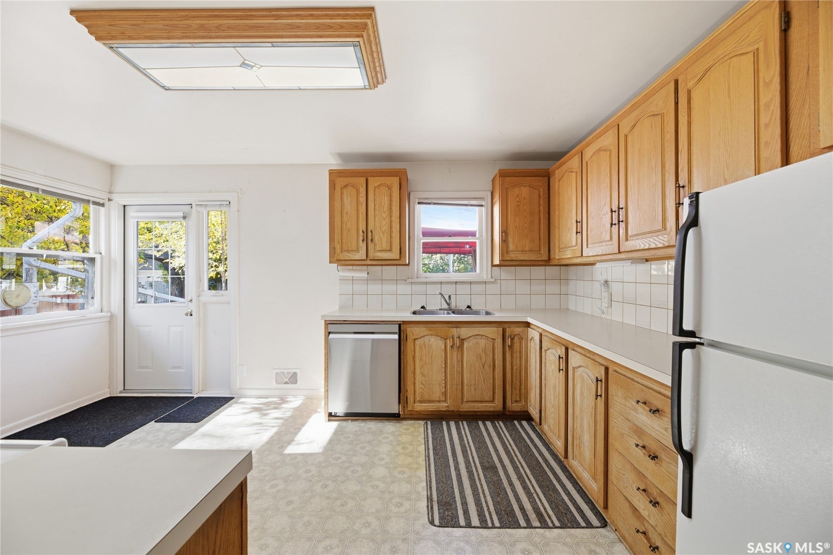 Wood-finish cabinetry, white tile backsplash, light-toned countertops, and a stainless steel dishwasher
