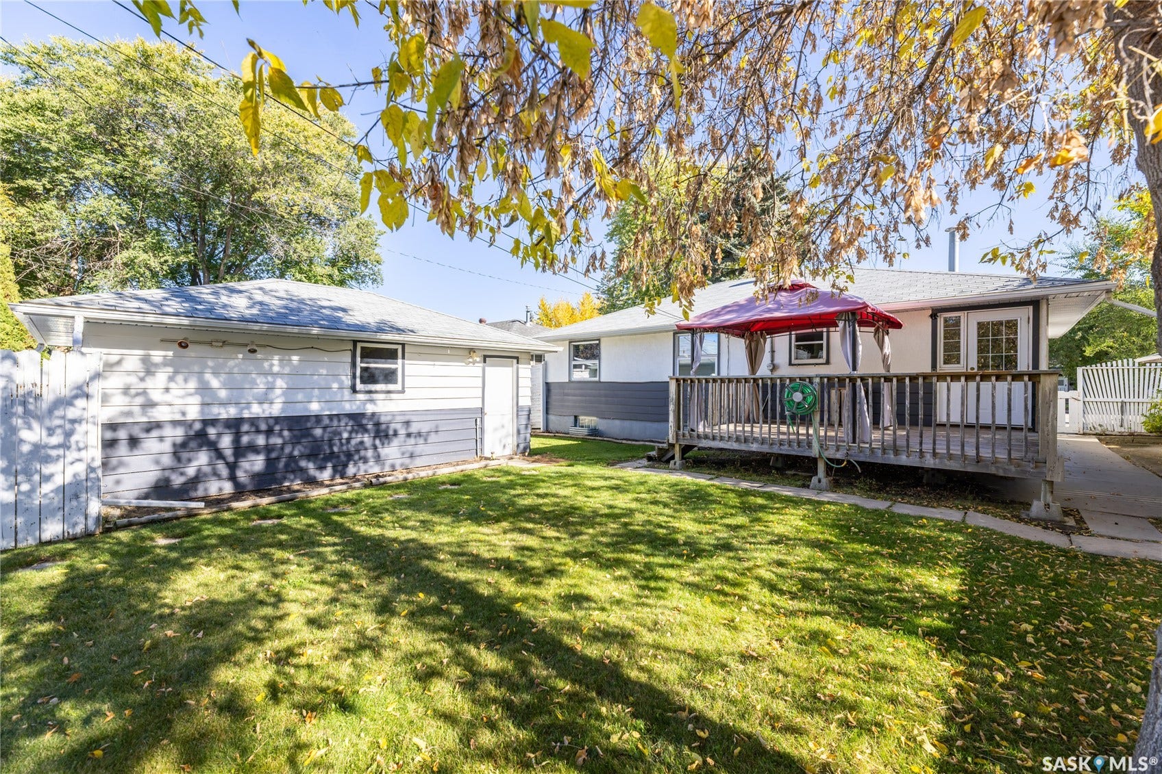 Expansive green lawn with mature trees, a multi-level wood deck, white picket fence, and a detached garage featuring a single window and side door