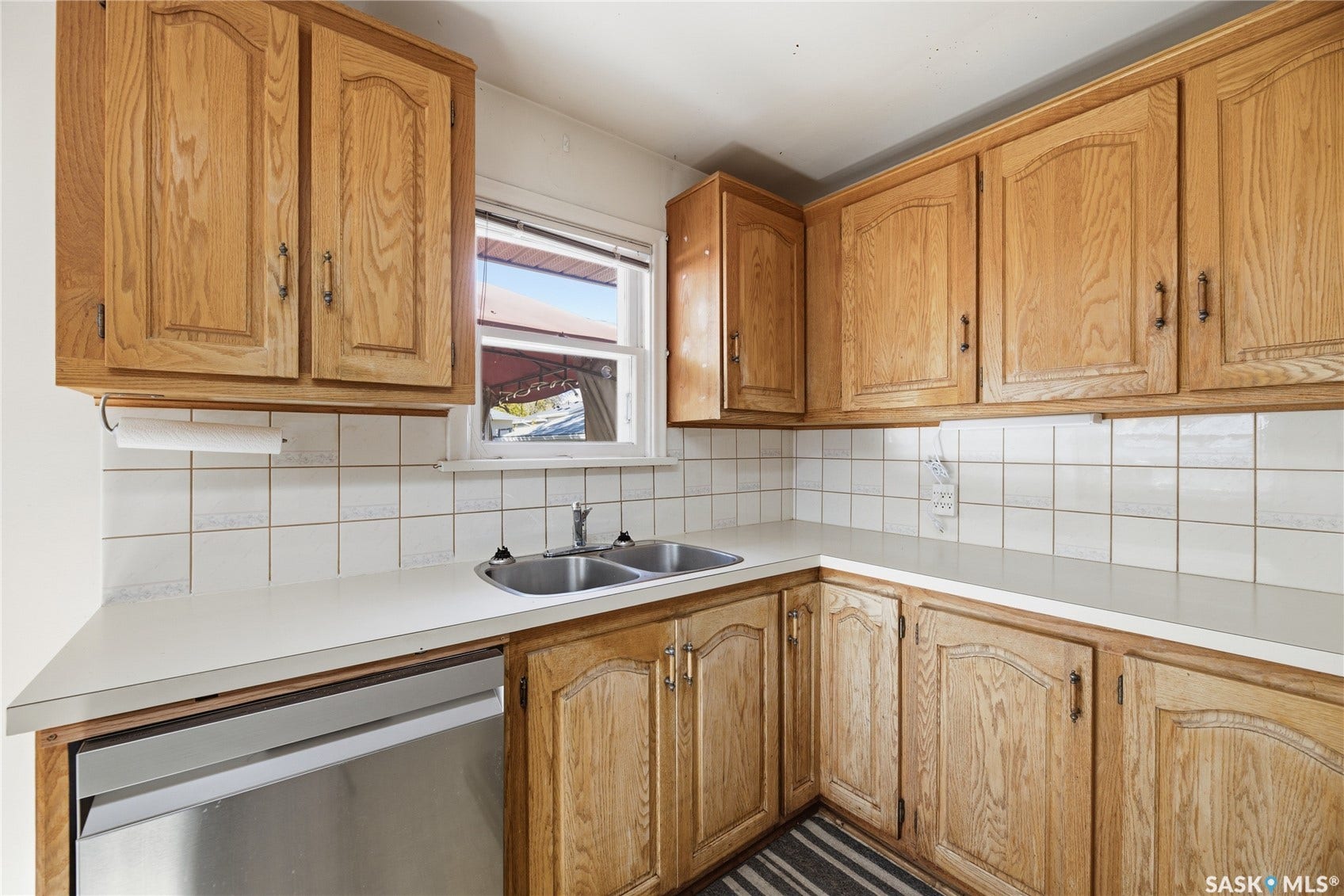Compact kitchen featuring warm wood cabinetry with paneled doors, laminate countertops, a stainless steel sink with single-lever faucet, white subway tile backsplash, and a window overlooking the exterior