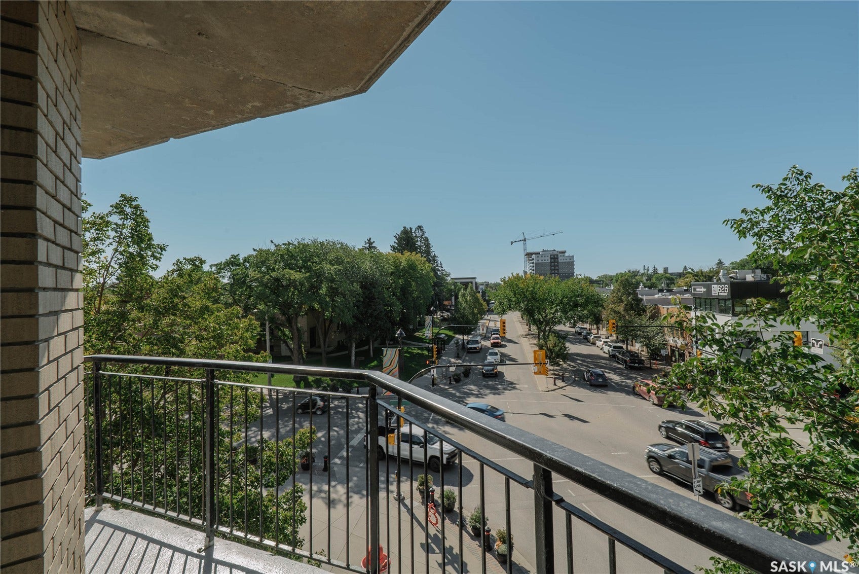 Brick exterior balcony with metal railing overlooking an urban streetscape featuring mature trees and commercial buildings