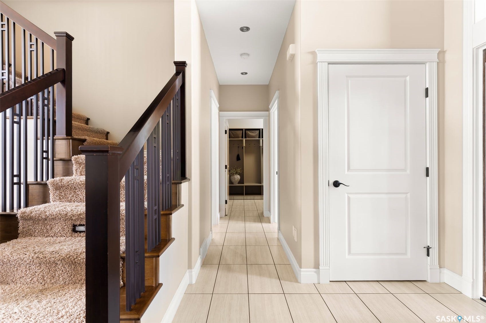 Staircase with plush carpet treads and dark wood newel posts, complemented by a tiled flooring hallway with recessed lighting and a paneled door