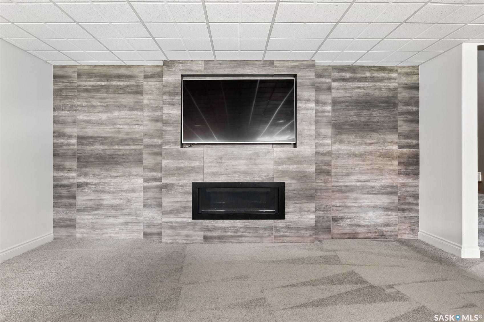 Feature wall with a built-in electric fireplace and mounted television, complemented by wall-to-wall carpeting and a grid-patterned ceiling