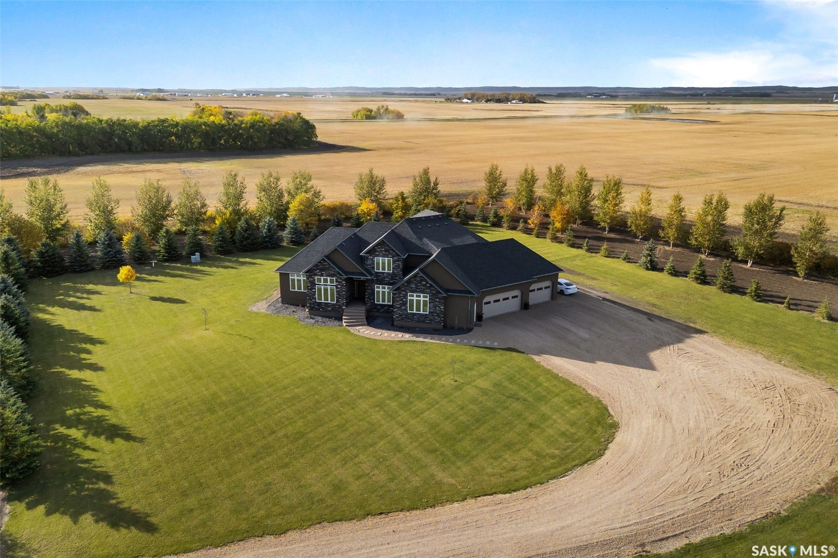Expansive property featuring a dark-toned residence with stone accents, a multi-car garage, a wide gravel driveway, and extensive green lawn areas