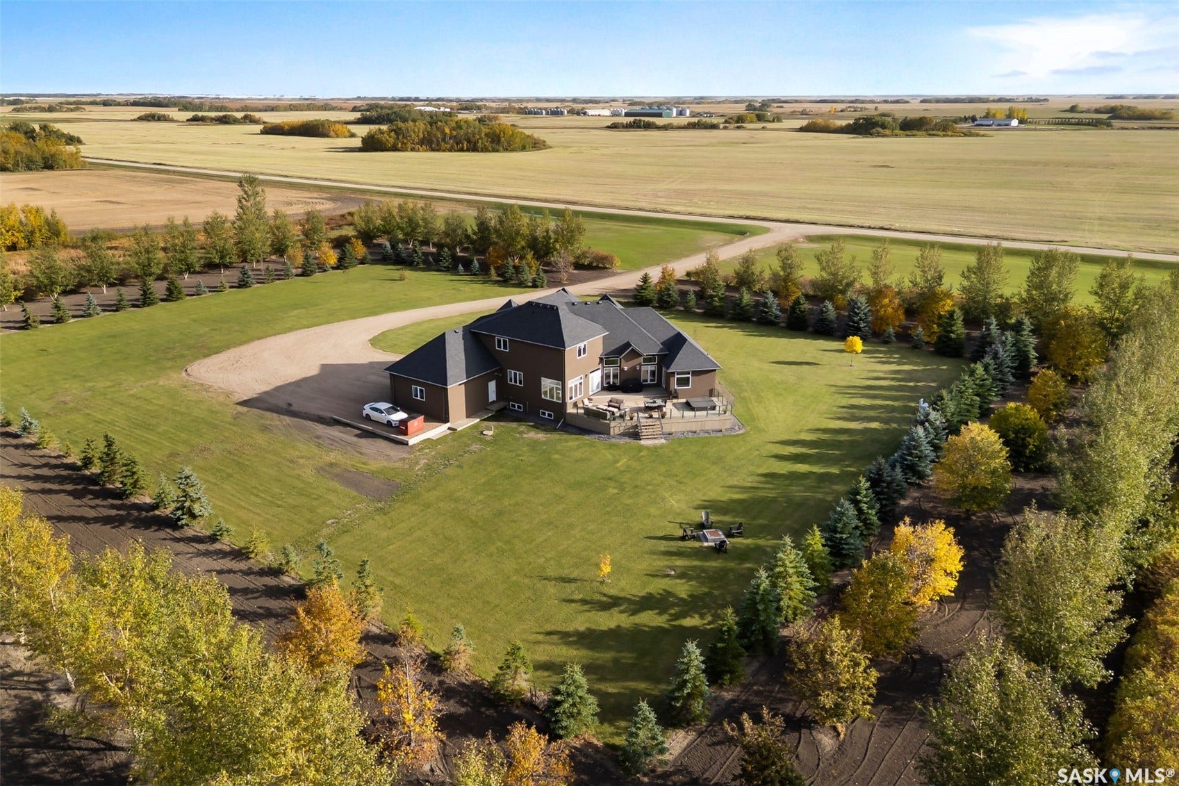 Expansive property featuring a brown exterior, dark shingle roof, a gravel driveway, and a spacious rear patio