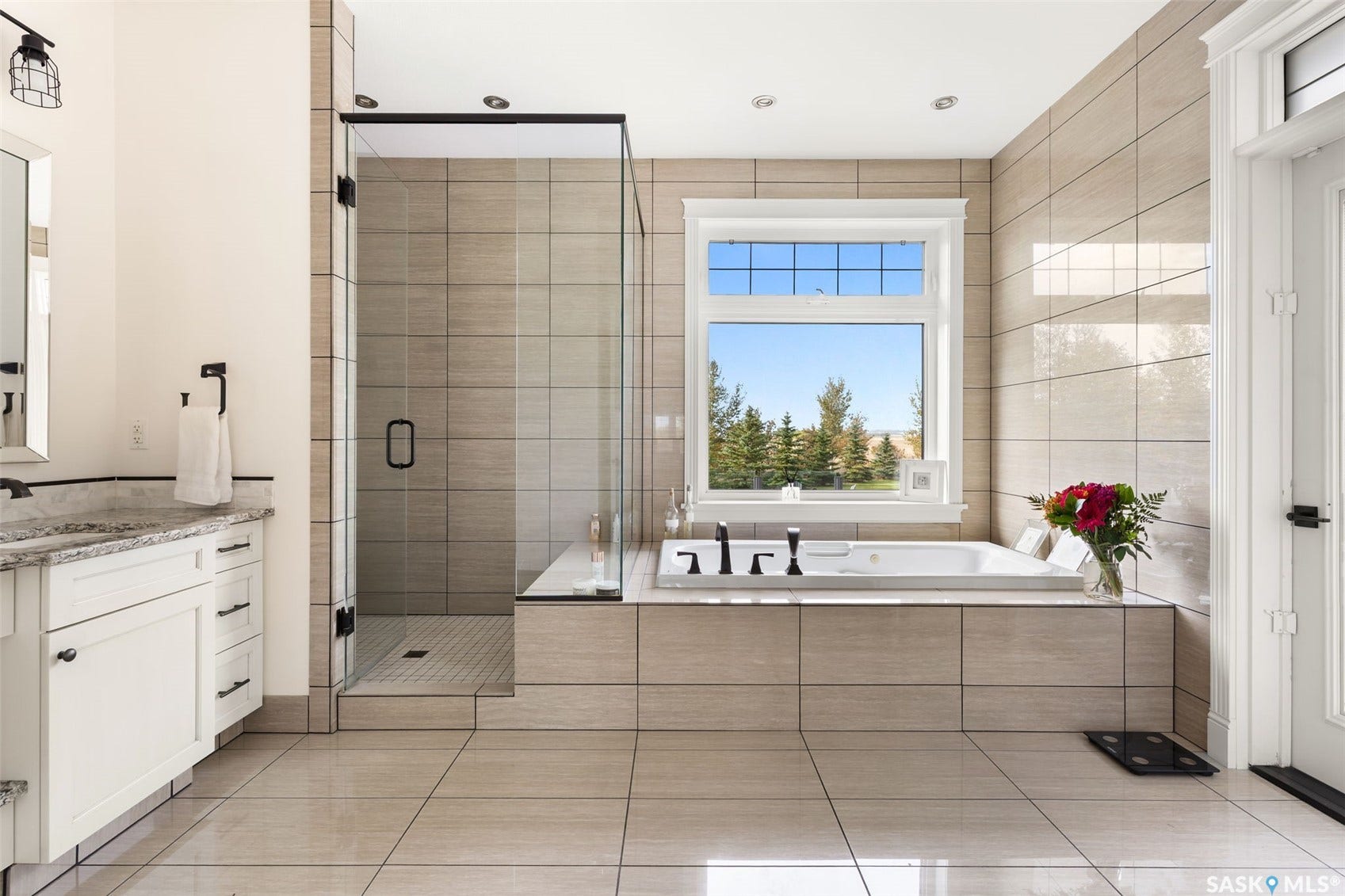 Spacious bathroom featuring a built-in soaking tub, a separate glass-enclosed shower, a vanity with stone-finish countertop, a large window, and tile flooring