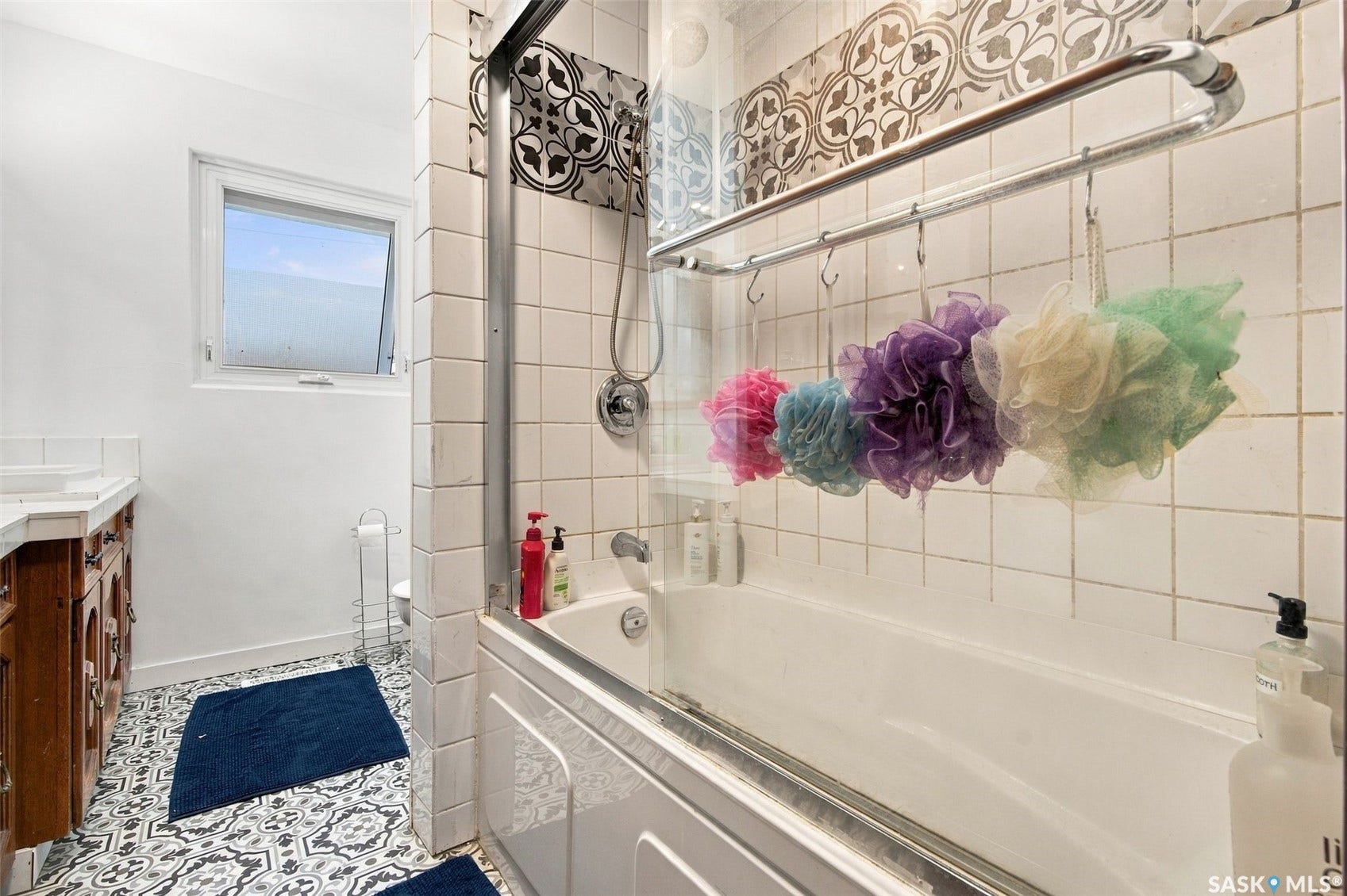 Bathtub with a glass enclosure, white subway tile surround, patterned accent tile, wood-finish vanity with a white countertop, and patterned floor tiling