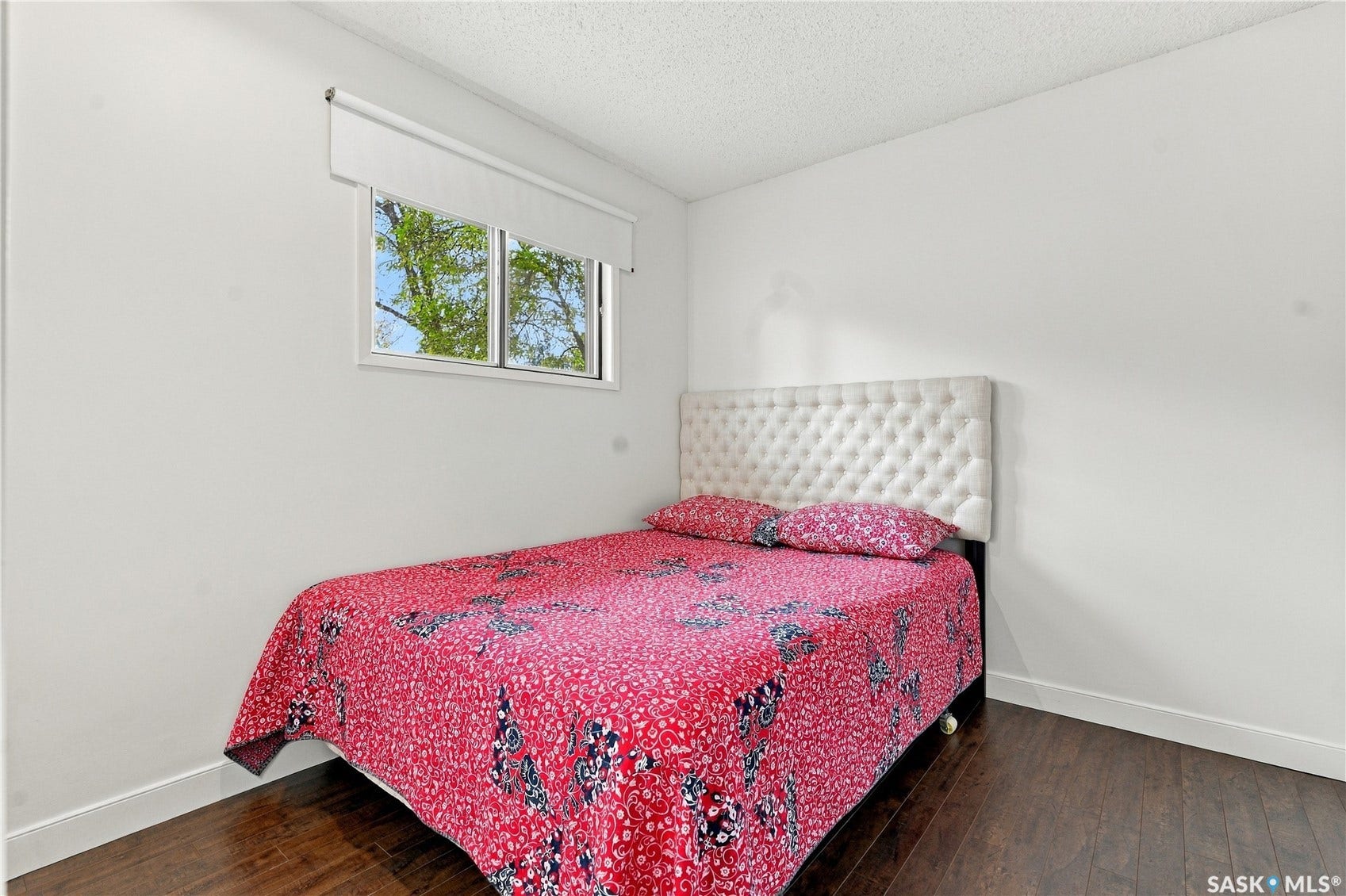 Window with white roller shade, dark wood-finish flooring, and white walls