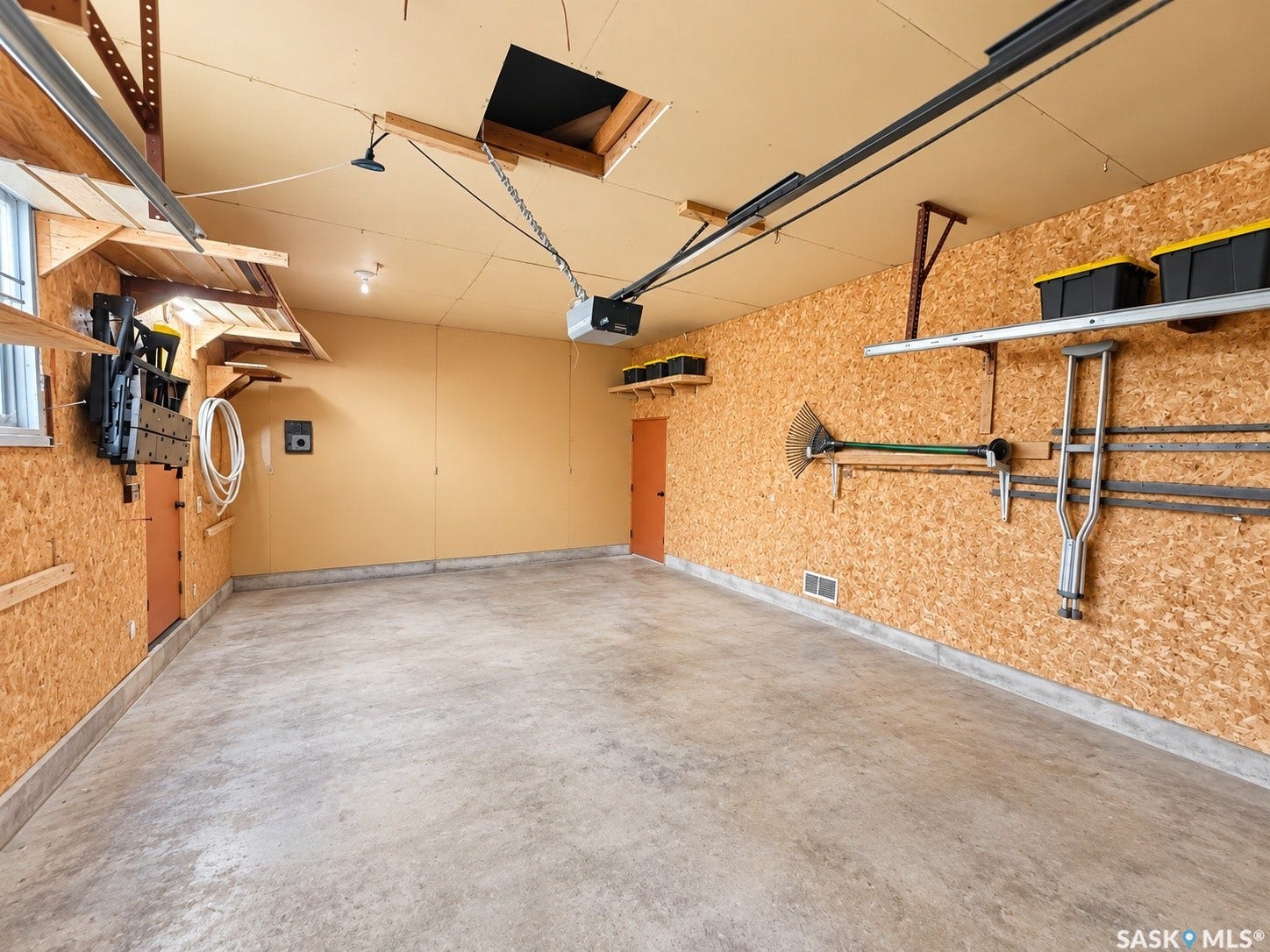5810 Sherwood Drive, Regina SK S4T 6Y7 Spacious garage with concrete flooring, exposed chipboard walls, utility shelving, and an overhead garage door opener system