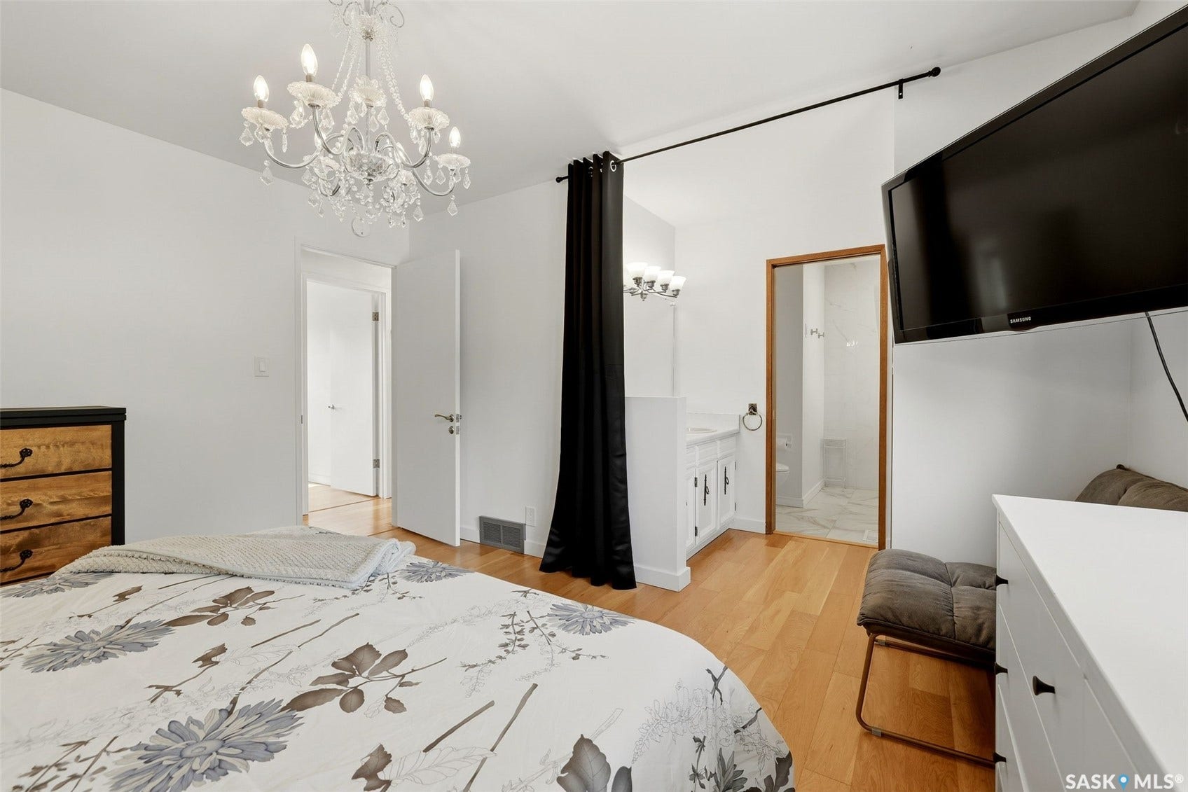 Wood-finish flooring, crystal chandelier, wall-mounted television, white vanity with wall-mounted lighting, and a white tiled shower enclosure