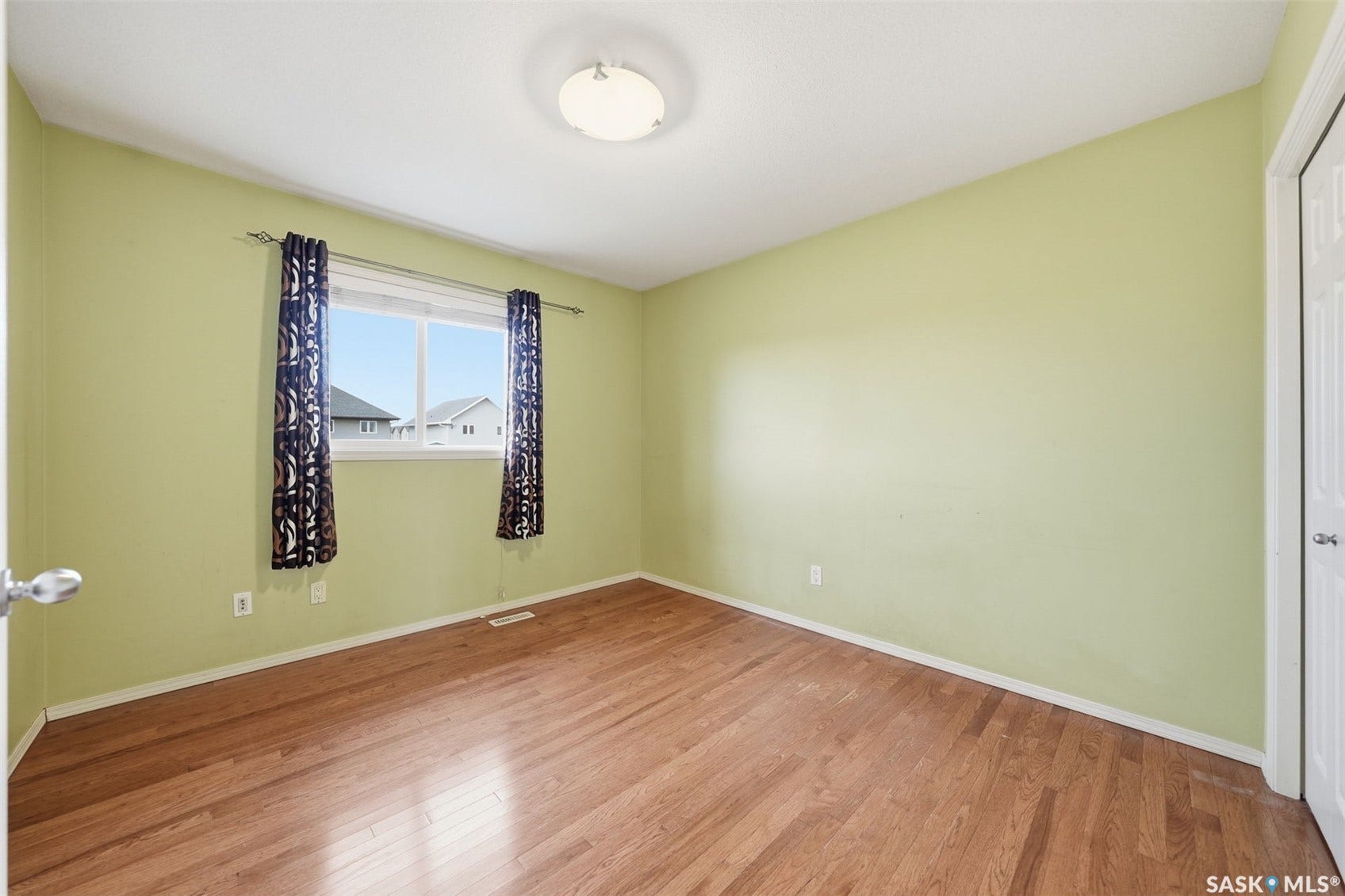 Bright and airy bedroom showcasing soft yellow walls, wood-finish flooring with a polished sheen, a large window with black patterned curtains allowing natural light, a ceiling-mounted light fixture, and a white interior door