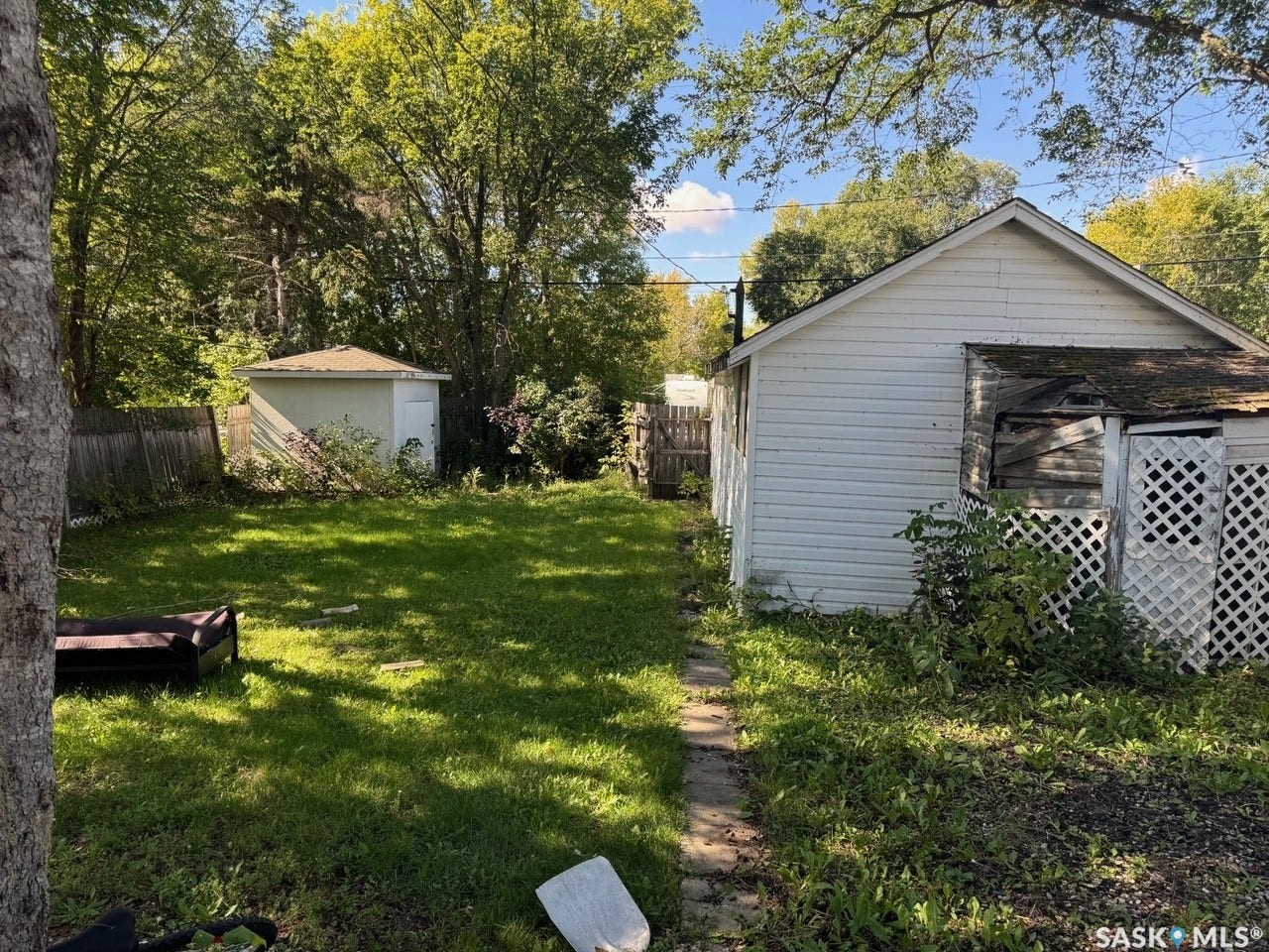 Spacious backyard featuring mature shade trees, lush lawn, and two detached outbuildings with siding and lattice detailing