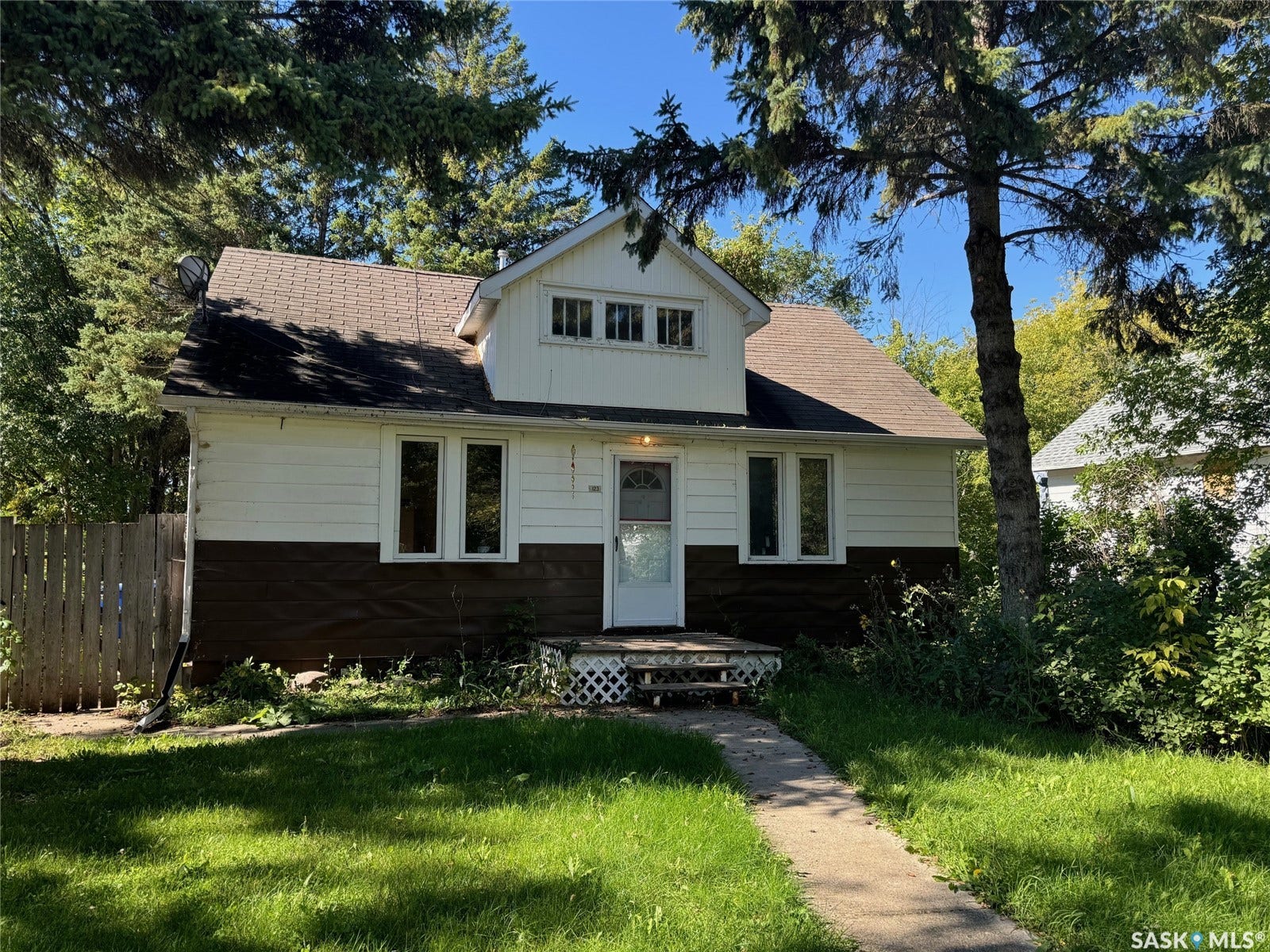 White and brown two-tone siding, asphalt shingle roof, front porch with lattice detailing, concrete walk, mature trees