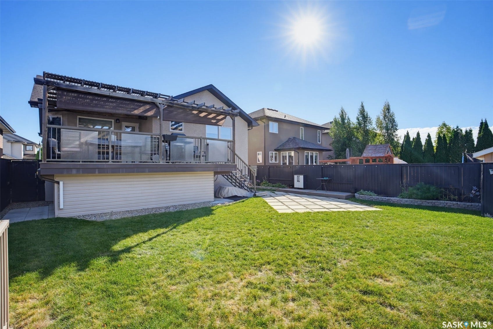 Expansive backyard featuring a manicured lawn, paved patio, elevated deck with glass railing, and a privacy fence