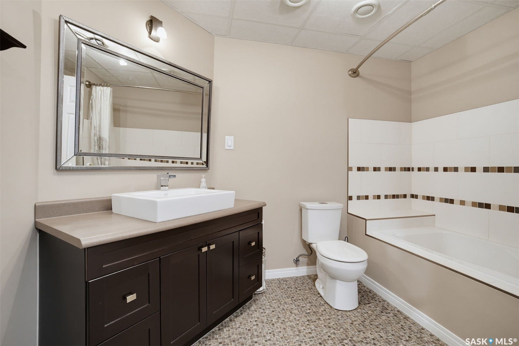Bathroom featuring a dark wood-finish vanity with a vessel sink, a rectangular framed mirror, pebble-finish flooring, and a tiled tub surround