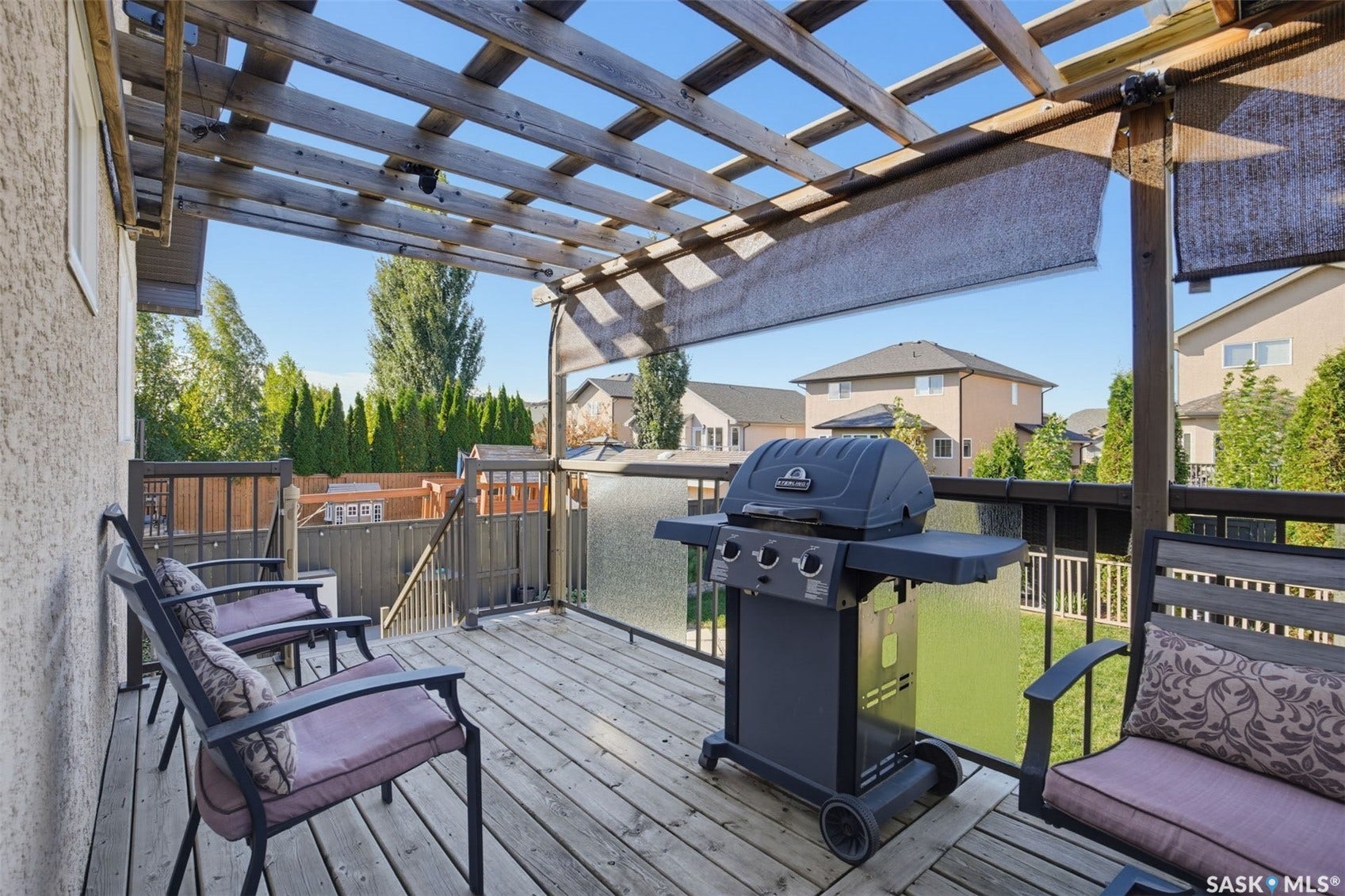 Outdoor wooden deck featuring a slatted pergola with retractable fabric shades, black metal railings with glass panels, and steps descending to the yard