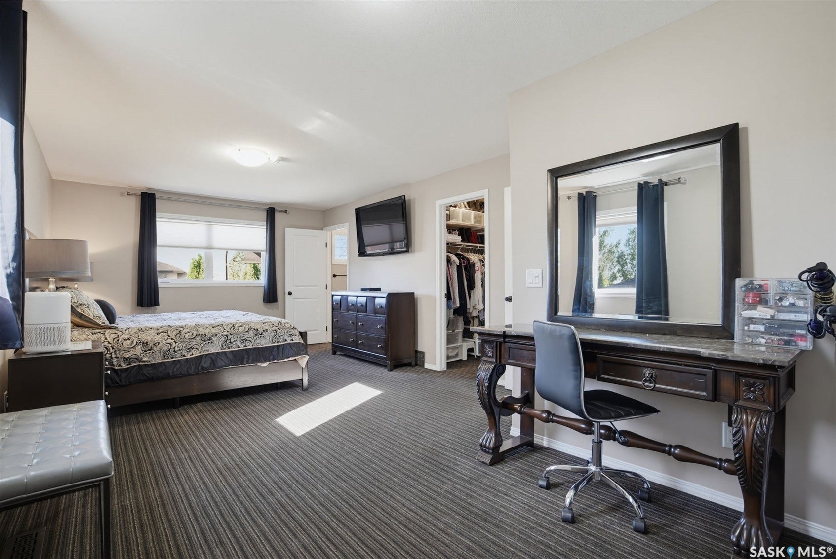 Spacious primary bedroom suite with dual windows providing natural light, striped wood-finish flooring throughout, and an en-suite vanity area with large framed mirror and desk workspace