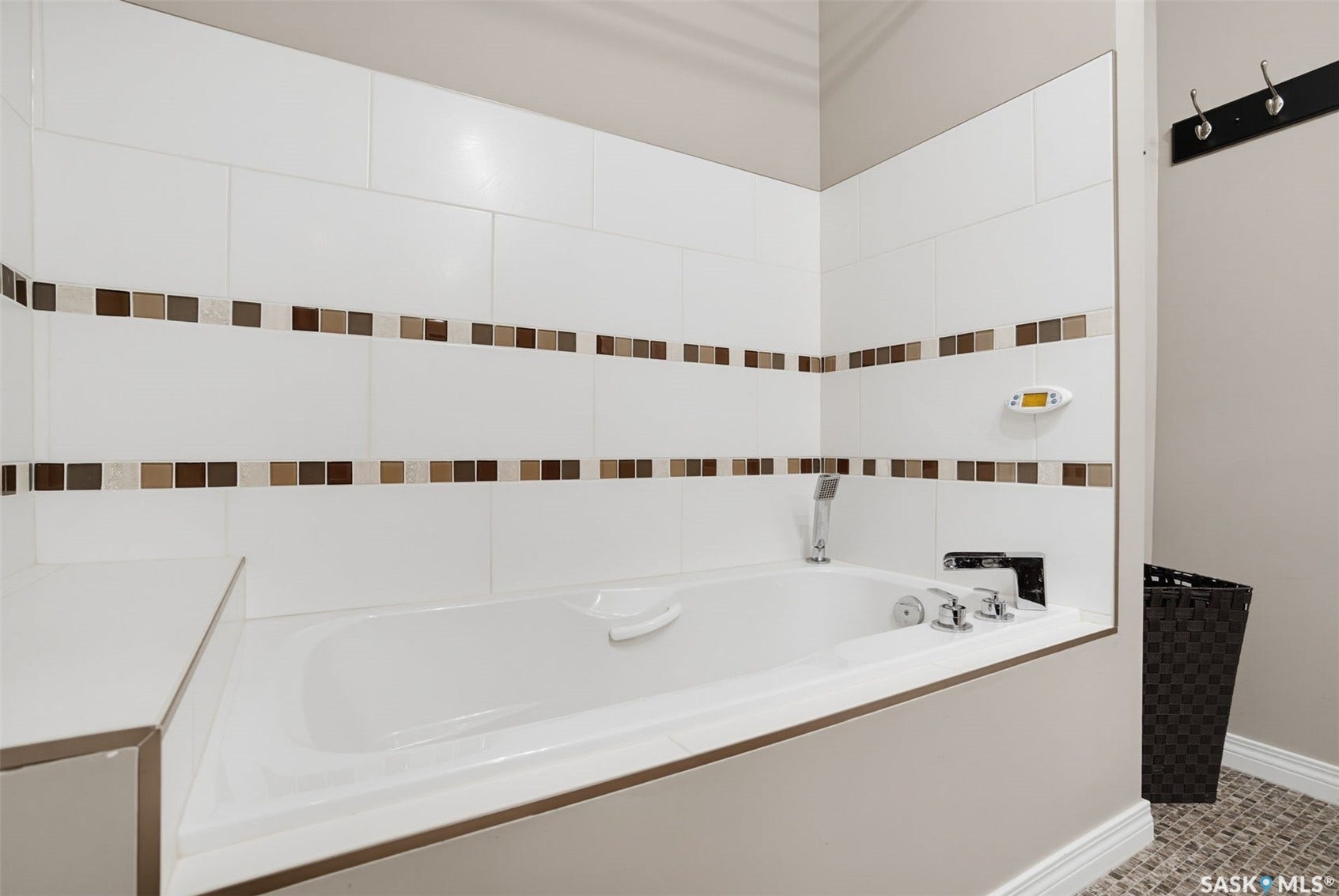 Built-in soaking tub with deck-mounted fixtures, white tile surround with mosaic accent, and textured floor tiling