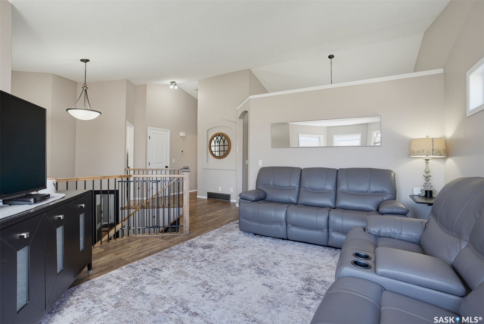 Vaulted ceiling with pendant lighting, wood-finish flooring, staircase with wood handrail and metal spindles, framed mirror, and arched entry with decorative window