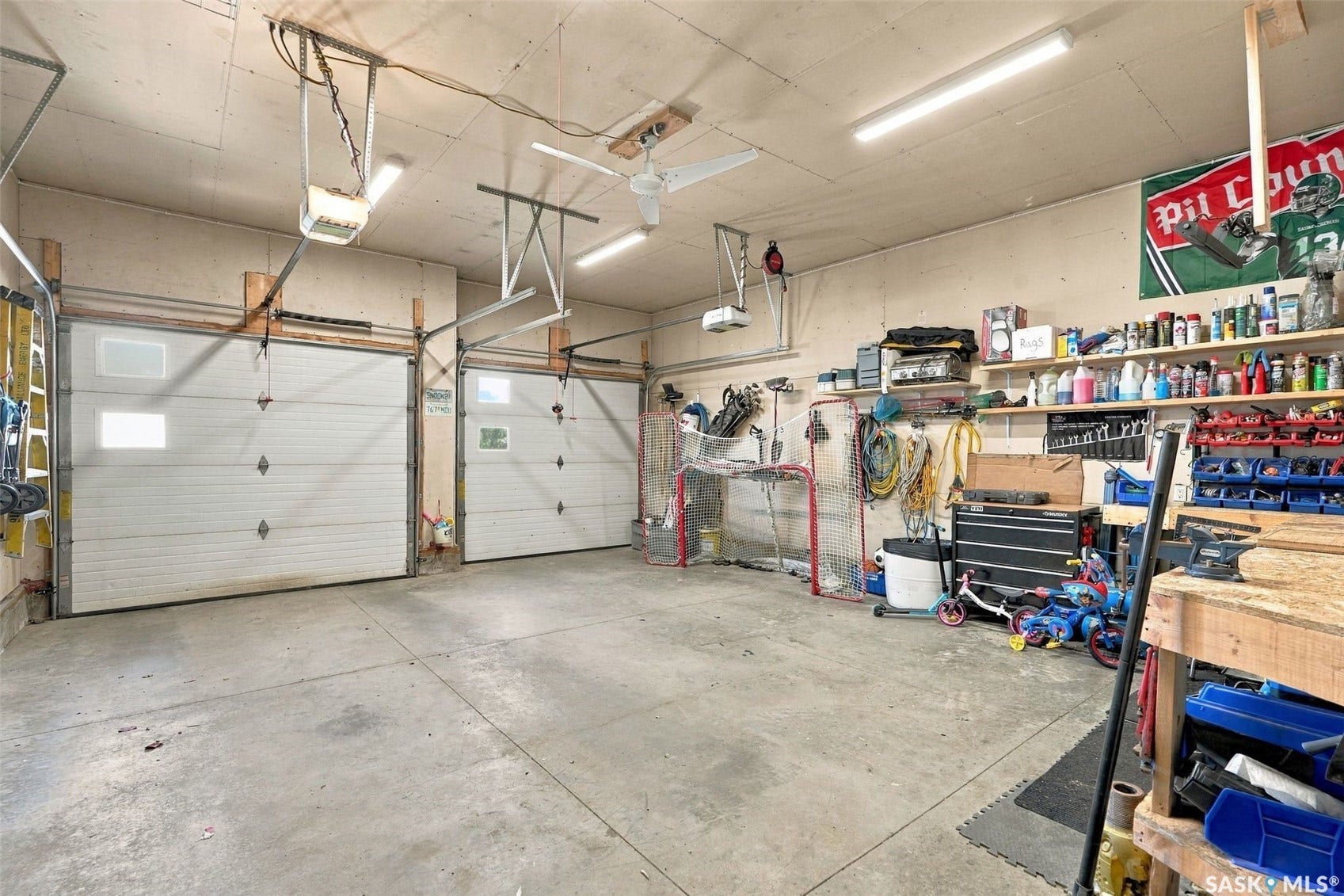Spacious two-car garage with concrete flooring, overhead storage systems, fluorescent lighting, and organized wall-mounted shelving