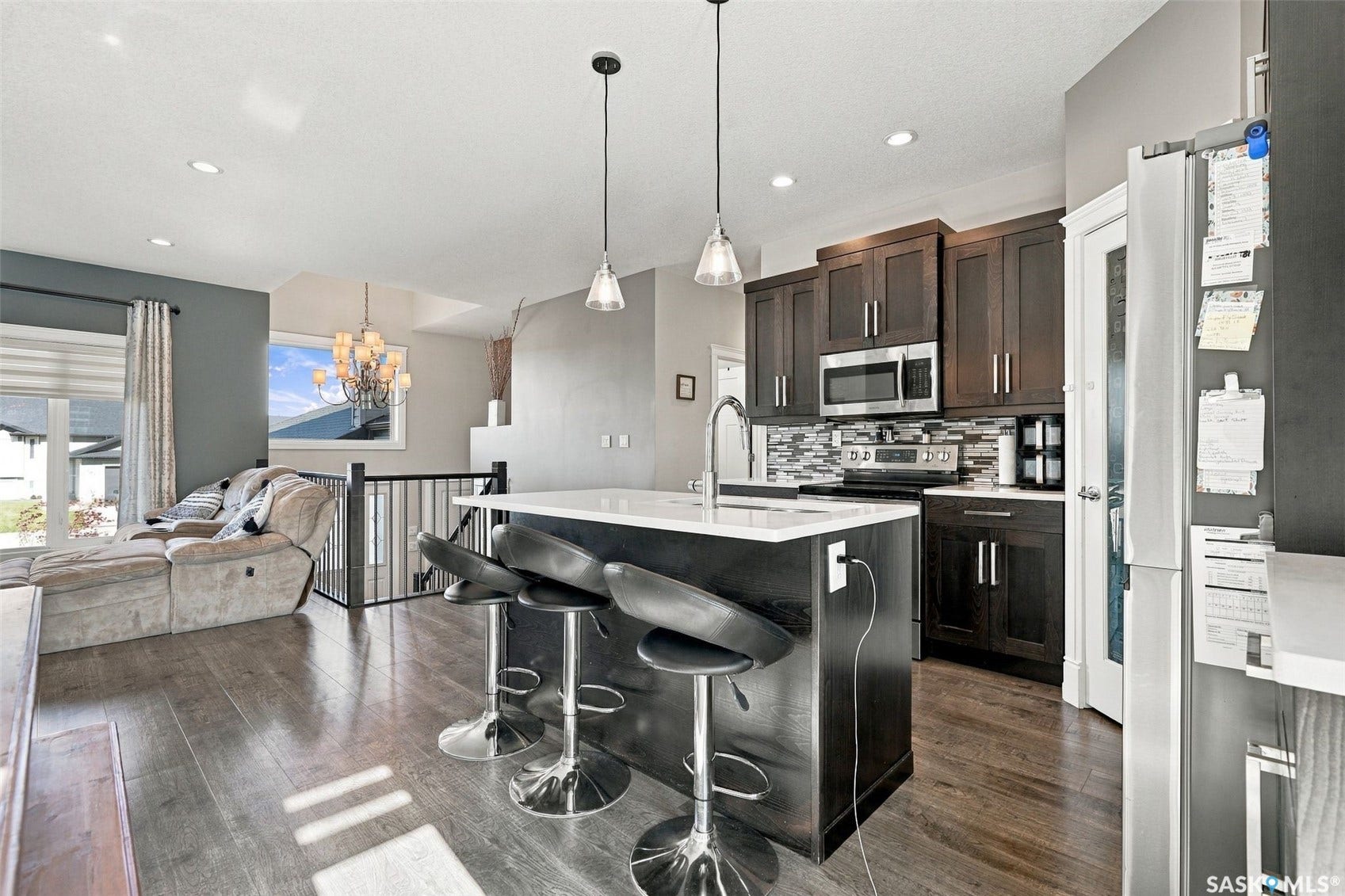 Open-concept kitchen and dining area featuring dark cabinetry with stainless steel appliances, a large waterfall-edge island with seating for multiple guests, white quartz countertops, and pendant lighting