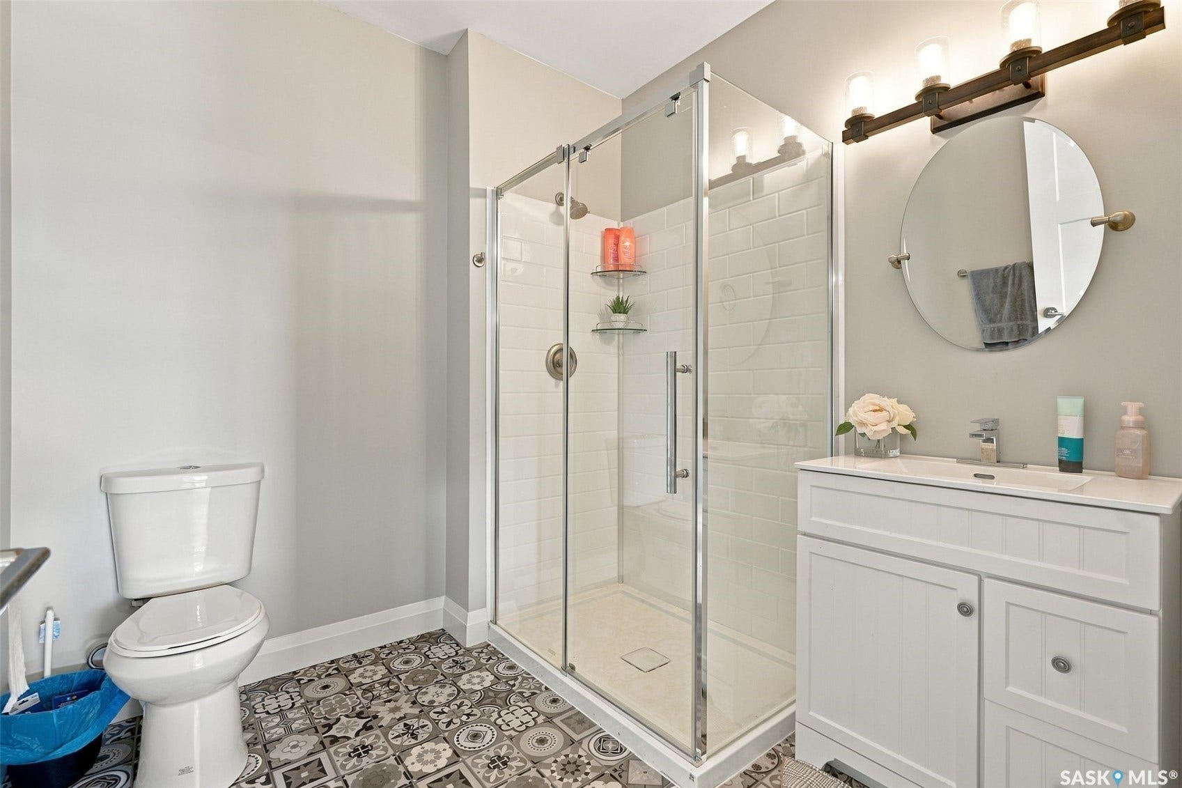 Glass-enclosed shower with subway tile surround, white vanity with integrated sink, patterned floor tile, and a round mirror