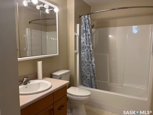 Bathroom featuring a circular sink with wood-finish vanity cabinetry, illuminated mirror with integrated lighting, ceramic toilet, and a bathtub/shower combination with subway tile surround and patterned shower curtain rod