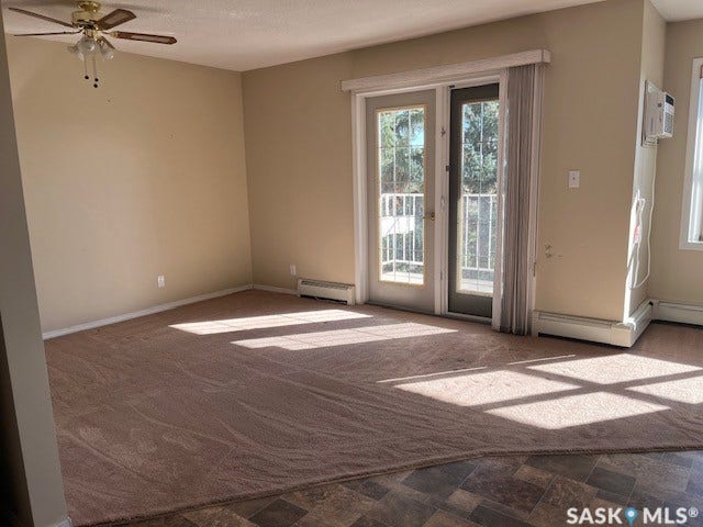 Textured carpeting, ceiling fan with integrated lighting, glass-paneled double doors, window-mounted air conditioning unit, and baseboard heating