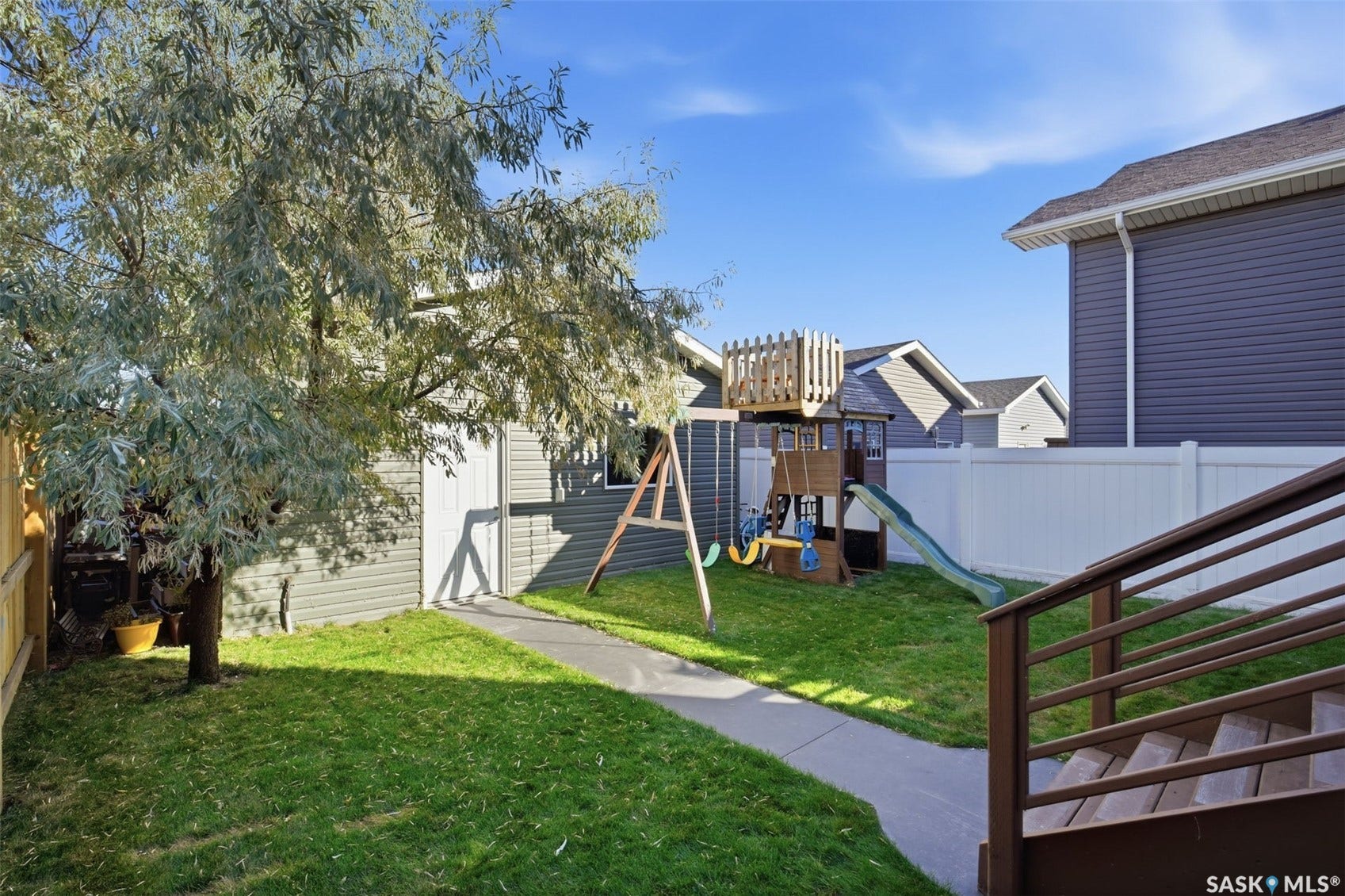 Expansive green lawn with a white privacy fence, mature tree, concrete pathway, and a built-in play structure featuring a slide and swings