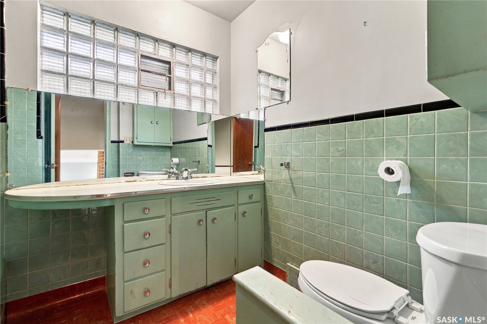 2727 Assiniboine Avenue, Regina SK S4S 1C7 Green tiled walls with black trim, glass block window, built-in vanity with integrated sink, full-width mirror, and a white toilet