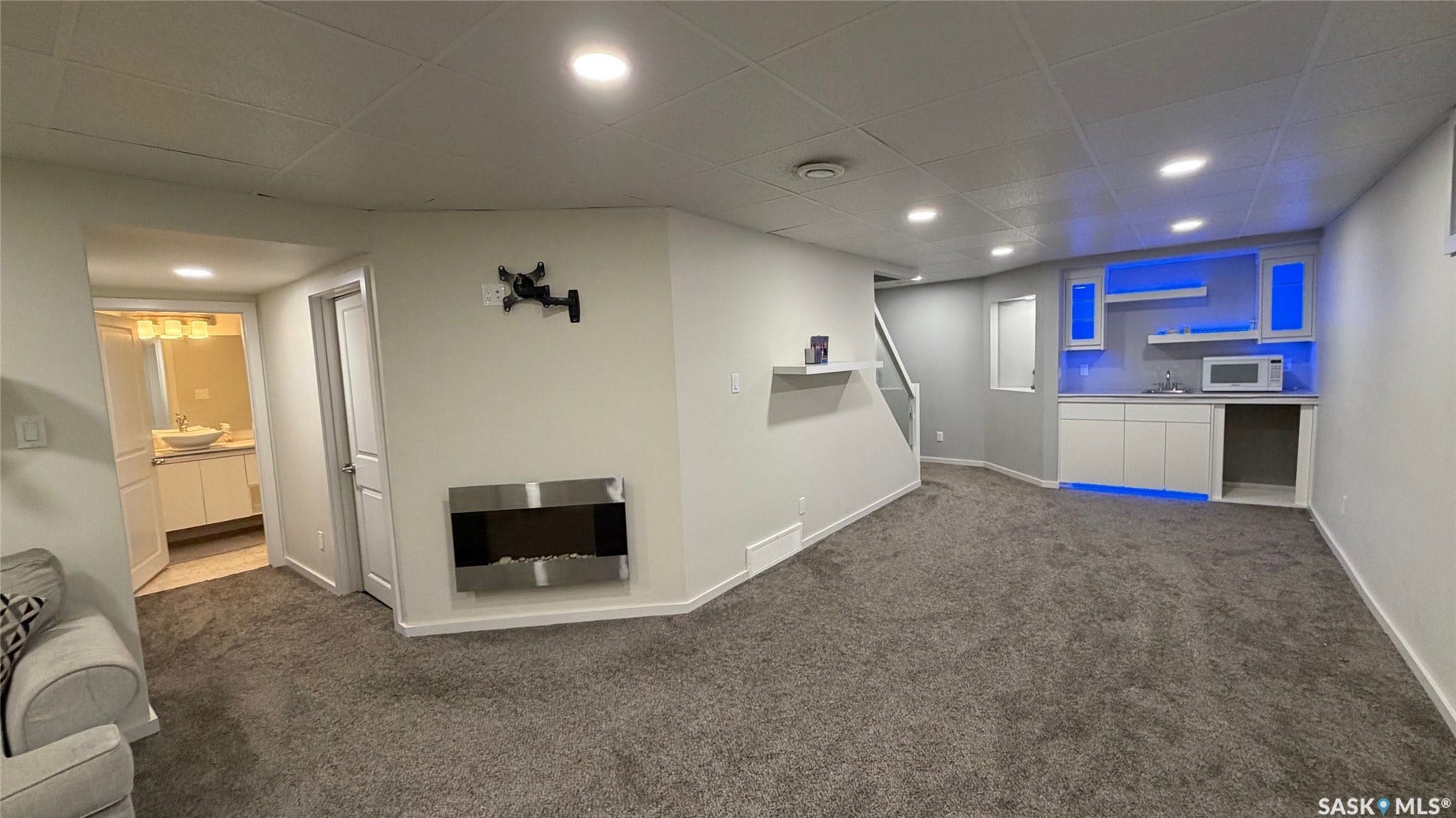 Recessed lighting throughout, gray carpet flooring, contemporary wall-mounted fireplace, white built-in cabinetry with under-cabinet lighting, and a visible bathroom vanity with a vessel sink