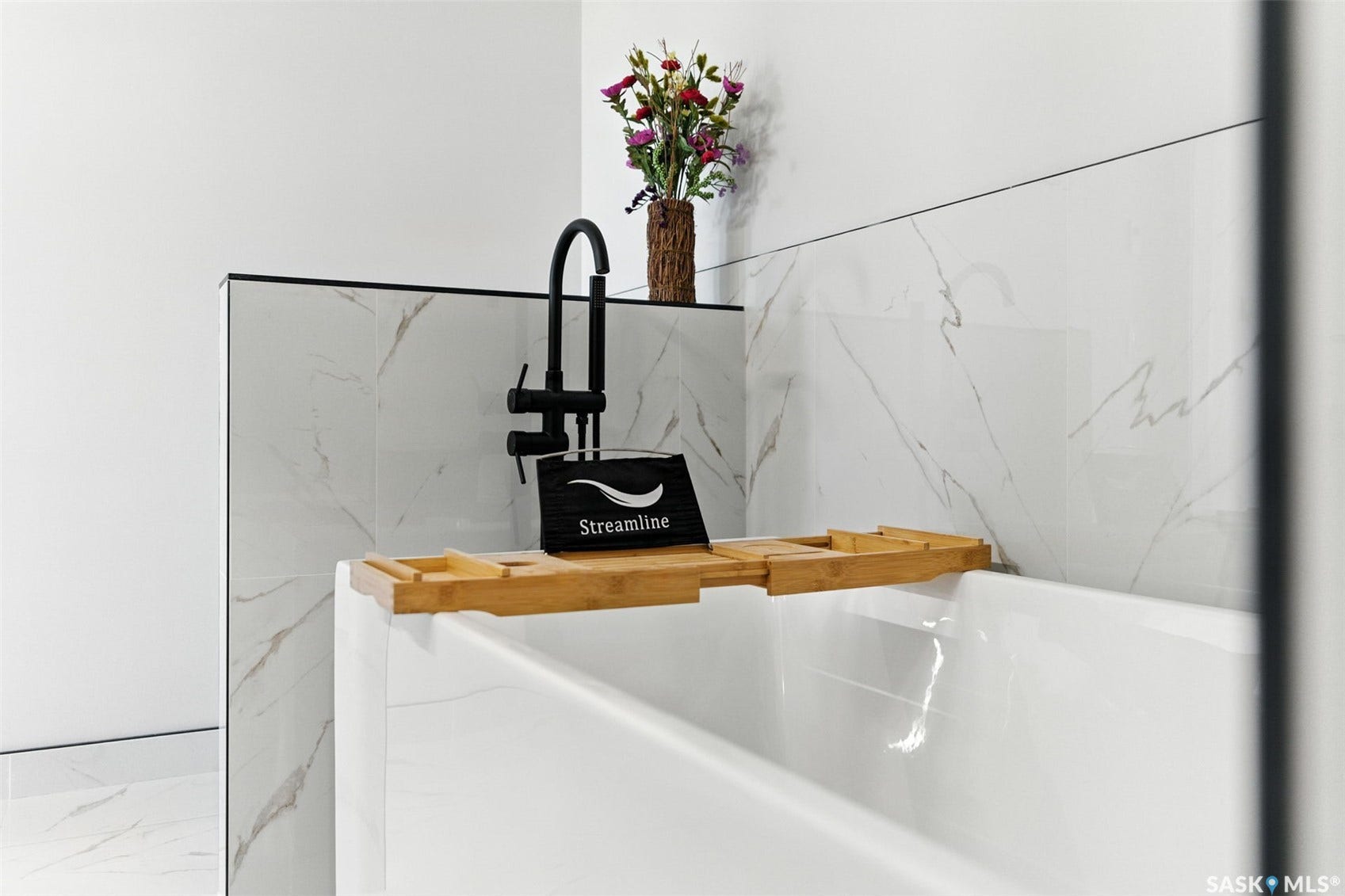 White soaking tub with a matte black wall-mounted faucet and white tile surround featuring gray veining