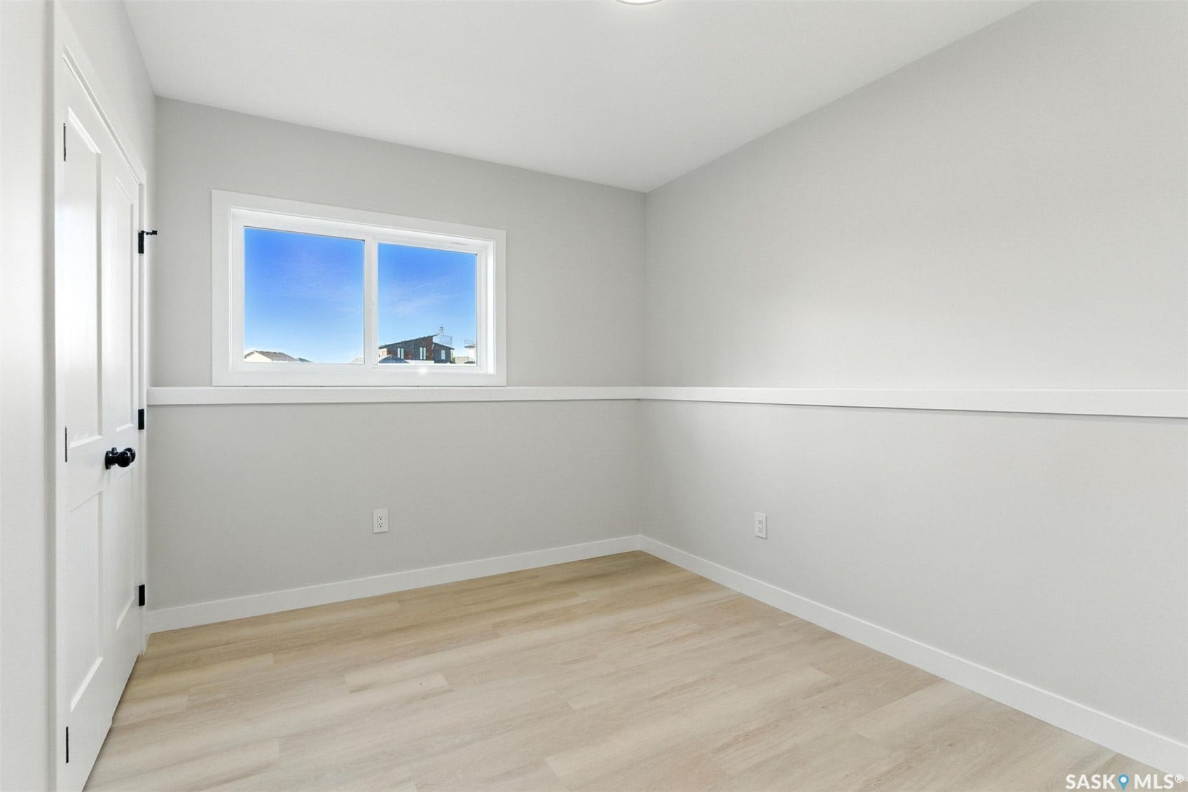 Wood-finish flooring, white trim, single window, white paneled door with black hardware, and light-colored wall paint