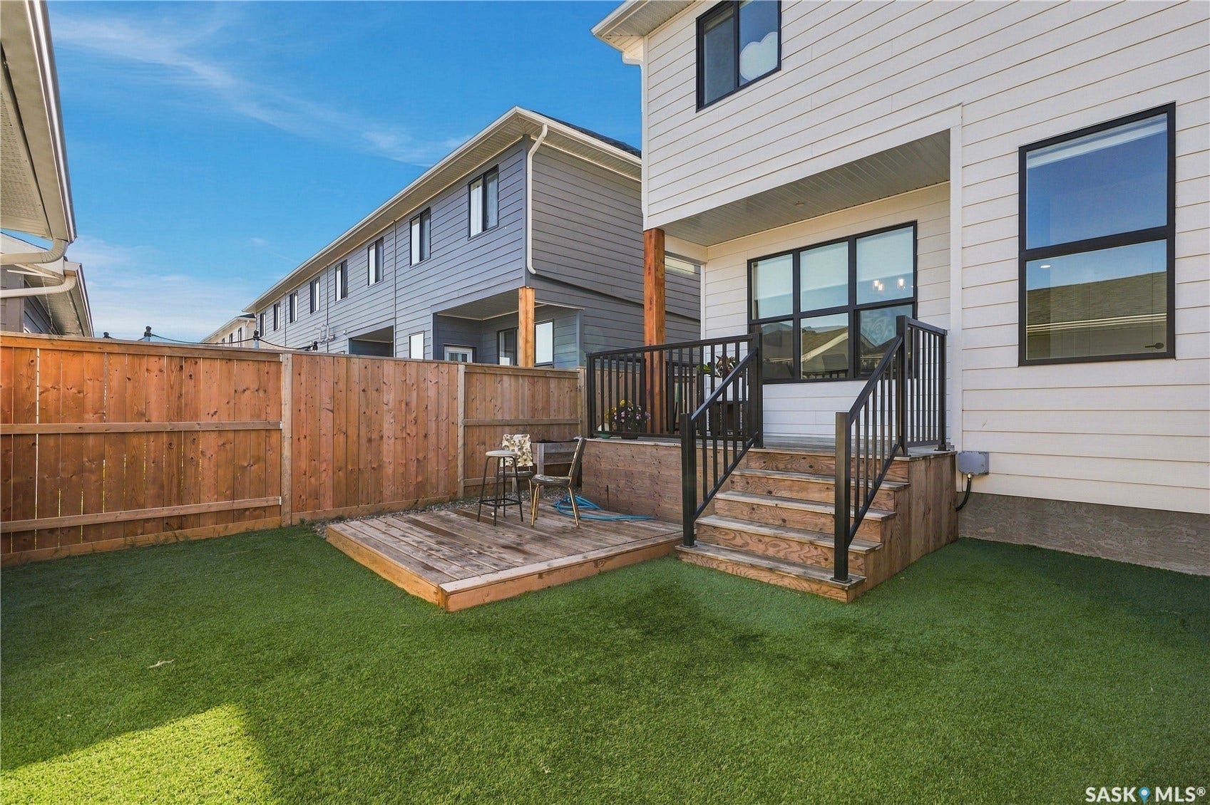 Rear exterior with white siding, elevated deck with black railings, wood stairs, fenced perimeter, and artificial turf
