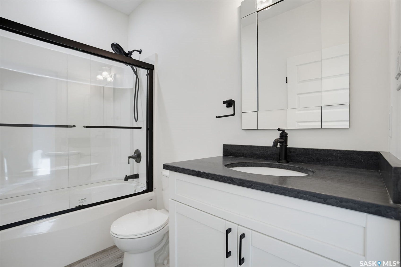 Built-in bathtub with glass sliding doors and matte black fixtures, white vanity with a dark countertop and integrated sink, and a mirrored medicine cabinet